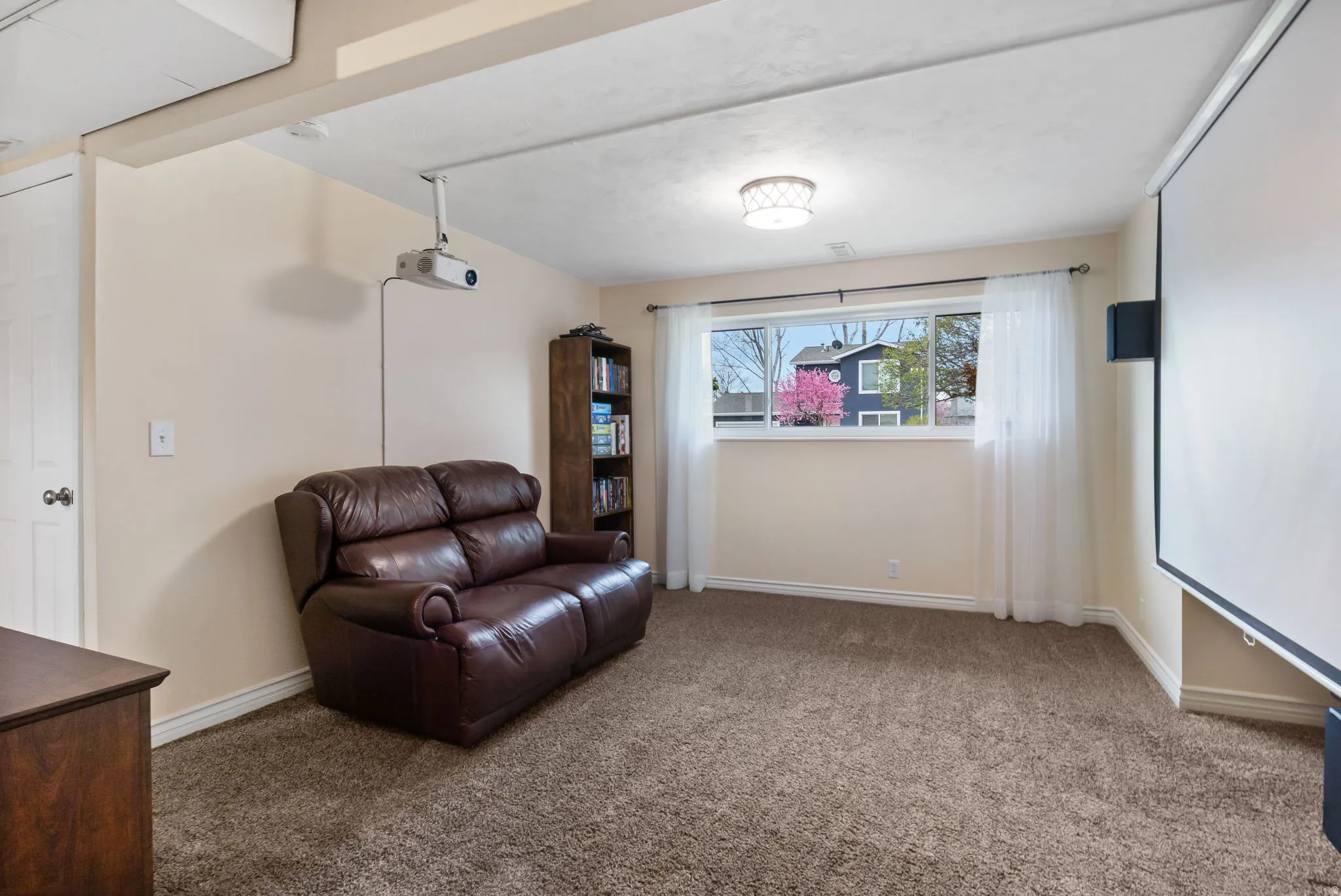 Carpeted room featuring a ceiling-mounted projector, large projection screen, built-in speaker, and a window with white sheers
