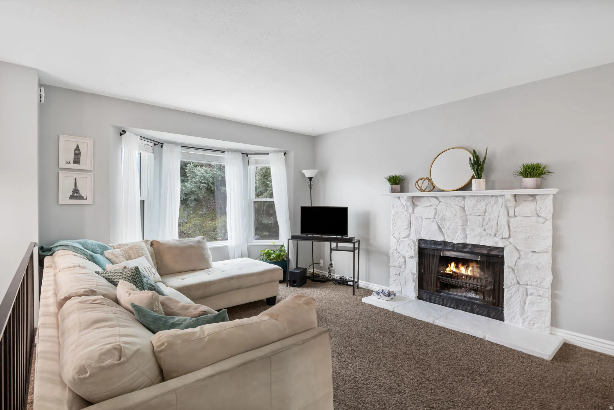 Living area featuring a stone-clad fireplace with hearth, a bay window with multiple panes, light gray wall paint, and brown carpeting
