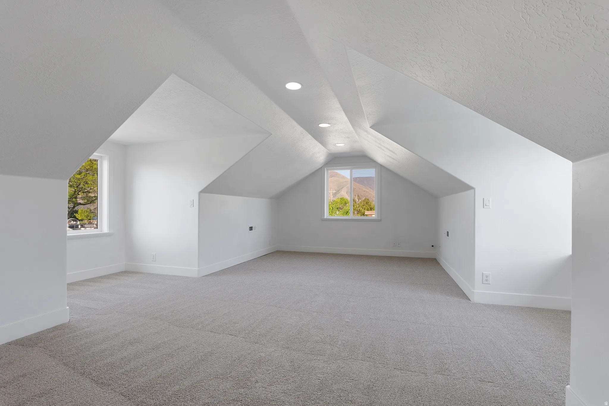 Spacious attic room featuring sloped ceilings, recessed lighting, light gray carpeting, crisp white walls, and multiple windows