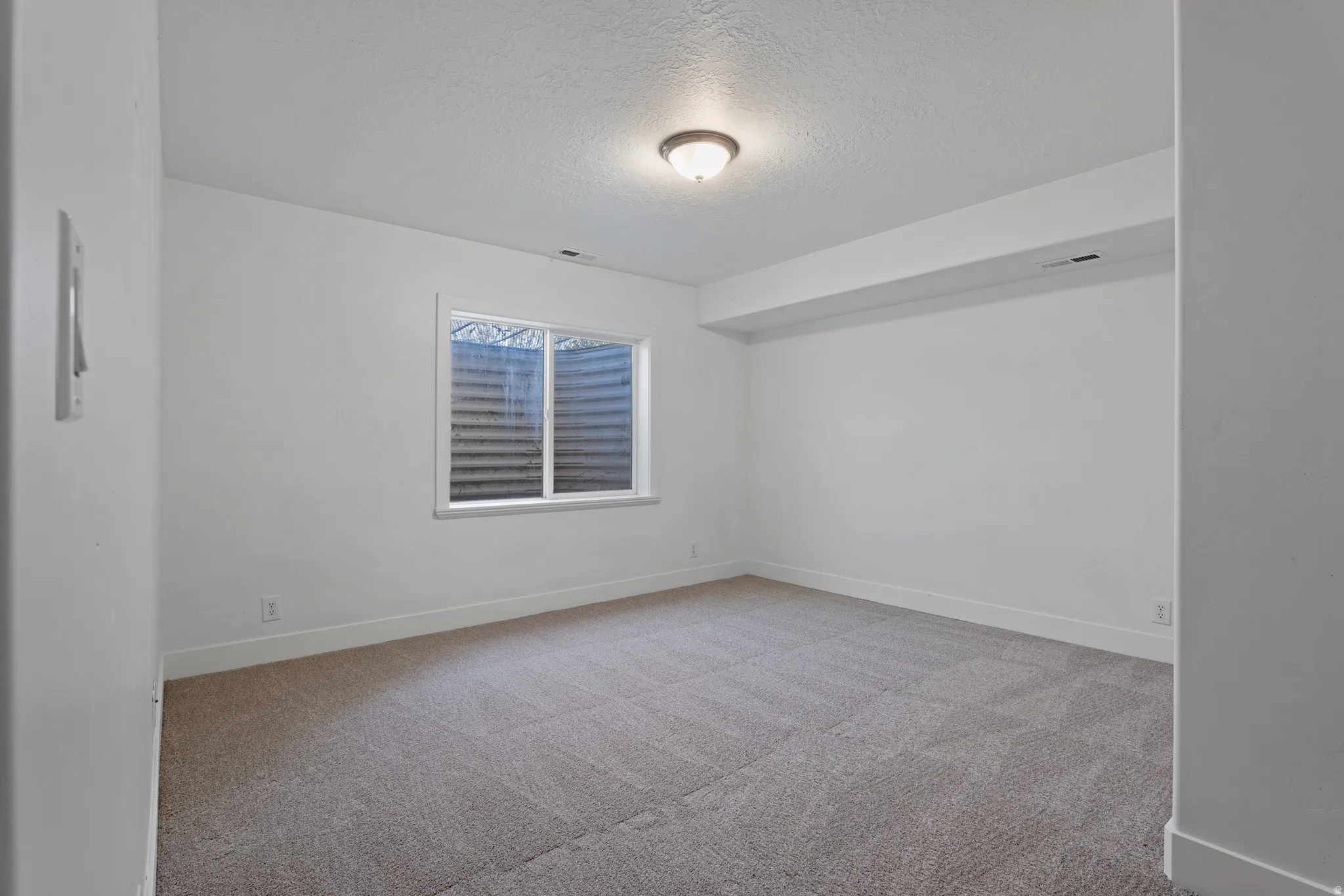 Spacious room featuring neutral carpeting, white painted walls, a white-framed window, a flush-mount ceiling light fixture, and white baseboards