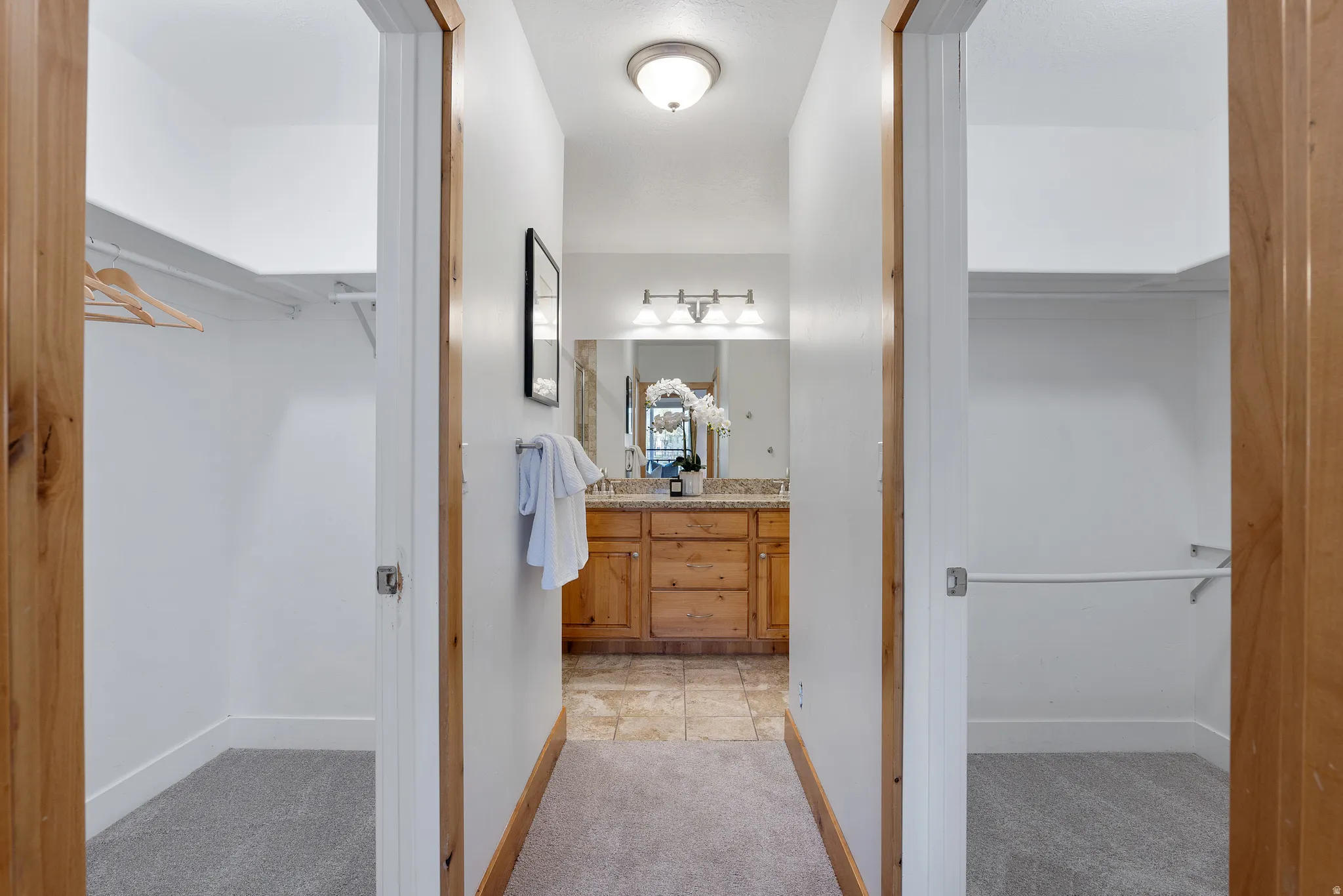 Hallway featuring two walk-in closets with carpet flooring, leading to a bathroom with a wood vanity, granite-finish countertop, and tile flooring