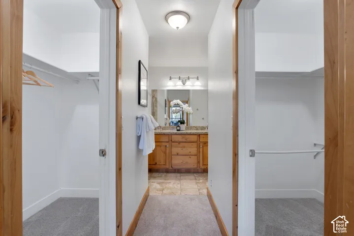 Hallway featuring two carpeted closets with shelving and hanging rods, leading to a bathroom with a dual-sink vanity, wood cabinetry, and a light-colored countertop
