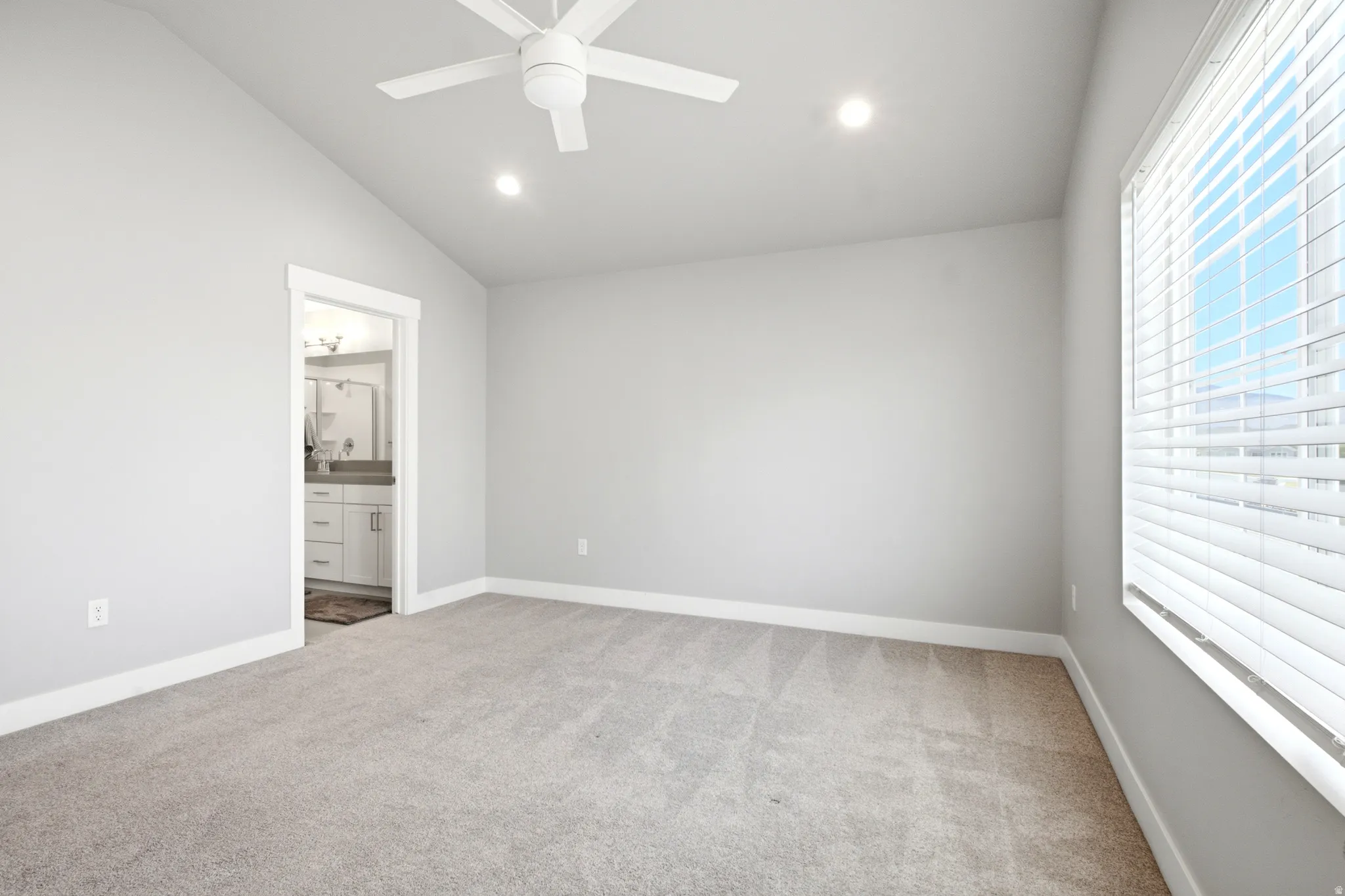 Spacious carpeted room featuring a white ceiling fan, recessed lighting, a large window with horizontal blinds, and a doorway revealing a bathroom with a double vanity and mirror
