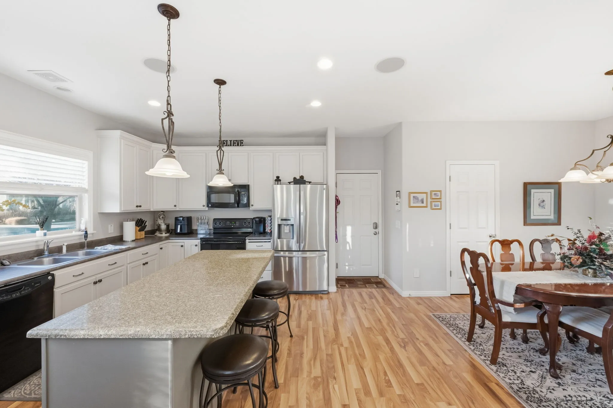 Kitchen featuring an elongated island with a light-toned countertop, white cabinetry with dark hardware, stainless steel refrigerator, black appliances, and pendant lighting