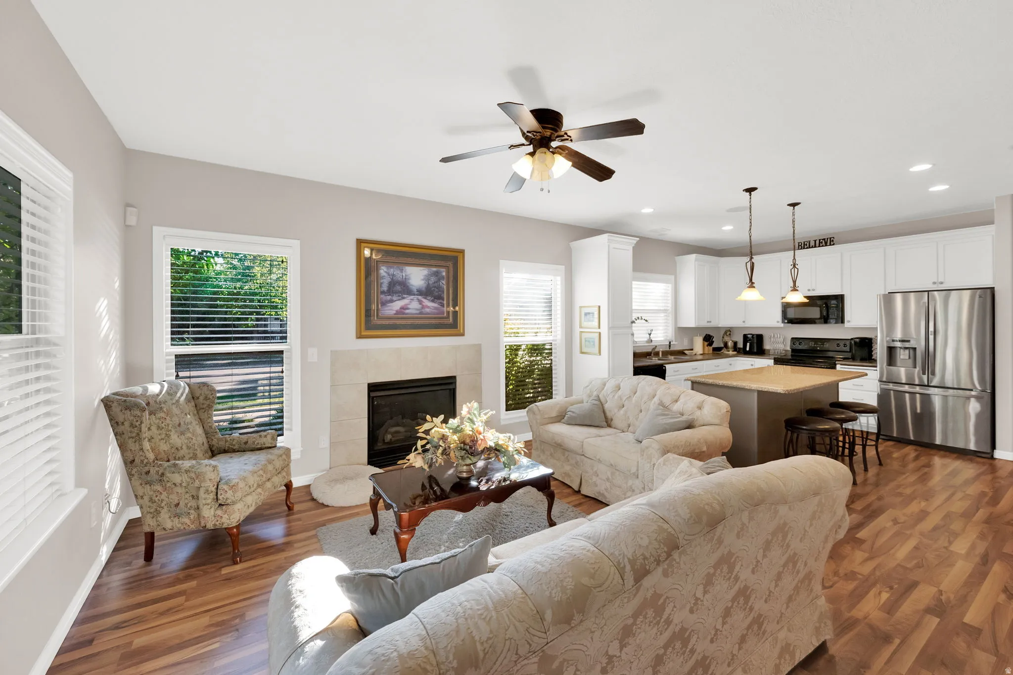 Open concept living area featuring wood-finish flooring, a tiled fireplace with mantel, and a ceiling fan with integrated lighting