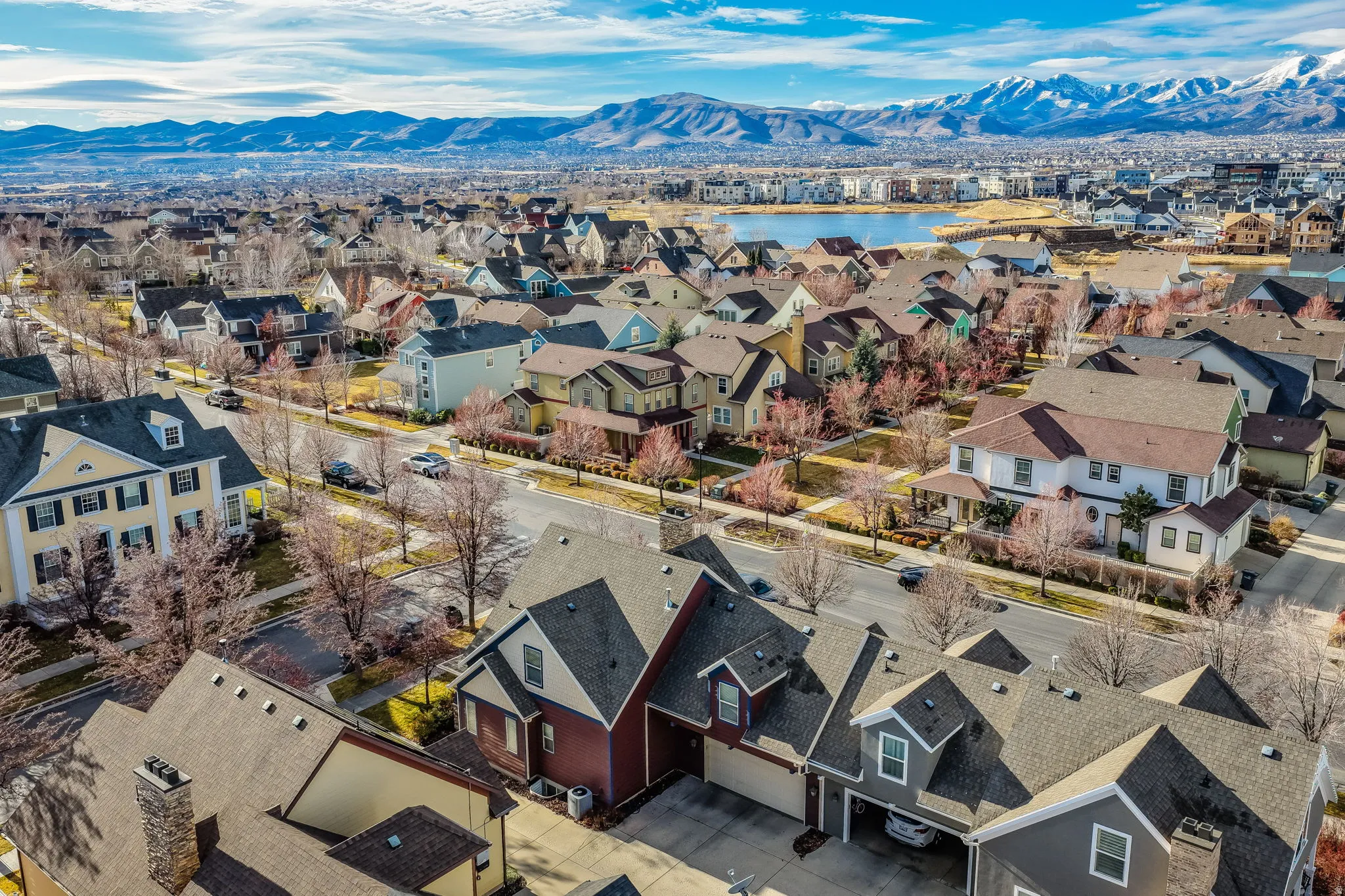 Expansive residential development featuring diverse architectural styles, visible roadways, mature tree-lined streets, a prominent body of water, and distant snow-capped mountains
