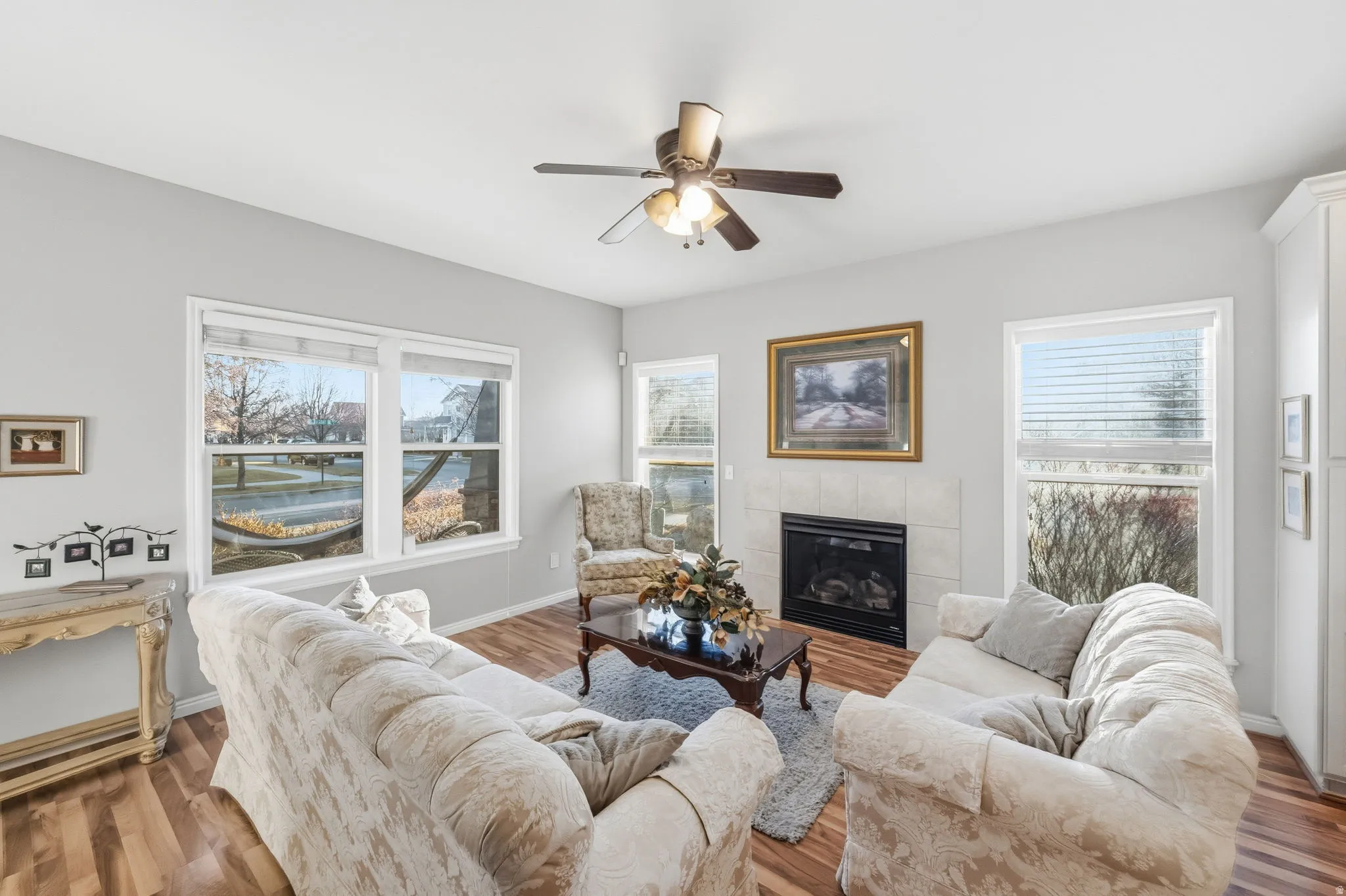 Spacious living area featuring wood-finish flooring, multiple windows, a ceiling fan, and a tiled fireplace