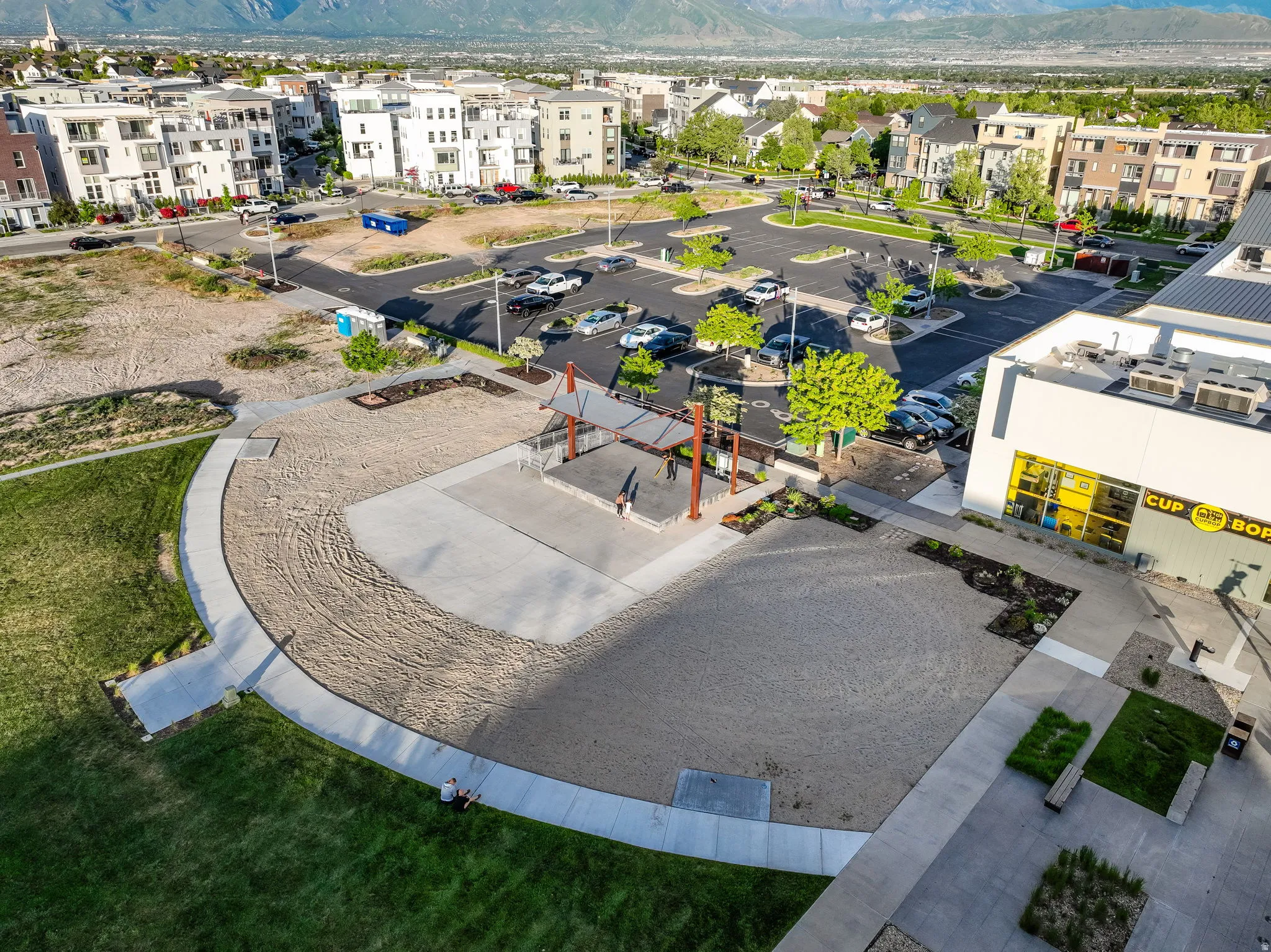 Community outdoor plaza featuring a shaded seating area, concrete pathways, and surrounding green spaces