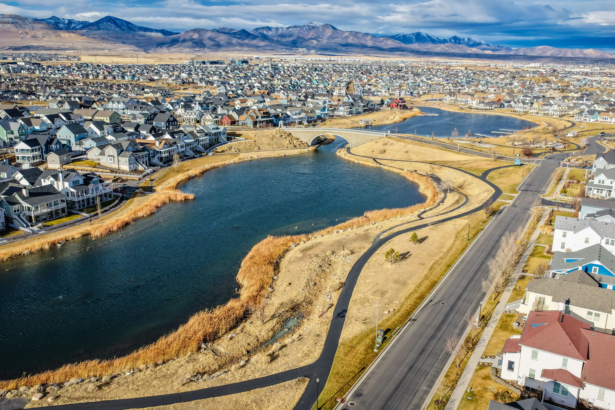 Expansive residential area featuring a large body of water, paved walking paths, a connecting bridge, and distant mountain ranges