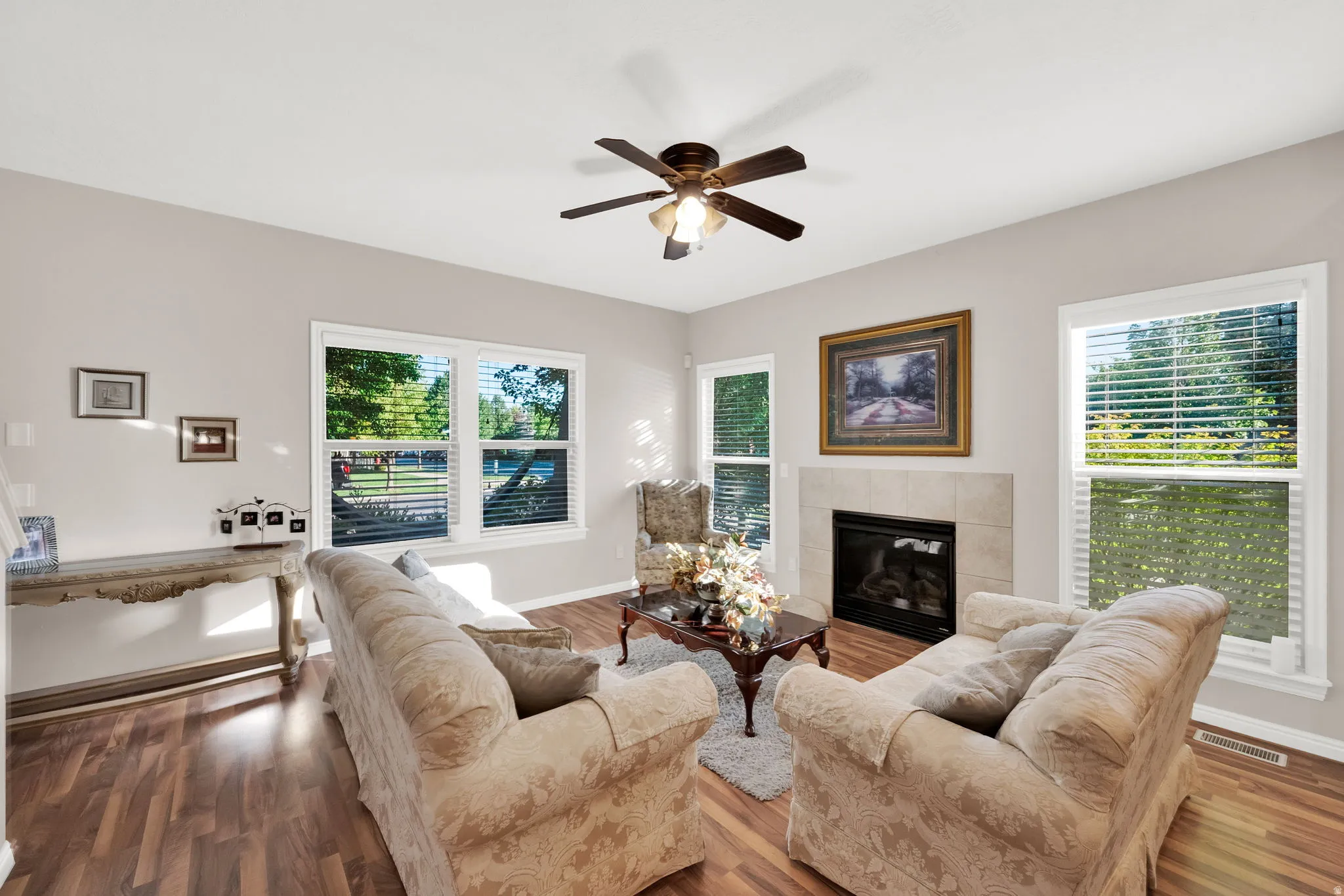 Living area featuring wood-finish flooring, a ceiling fan with integrated lighting, and multiple windows with blinds