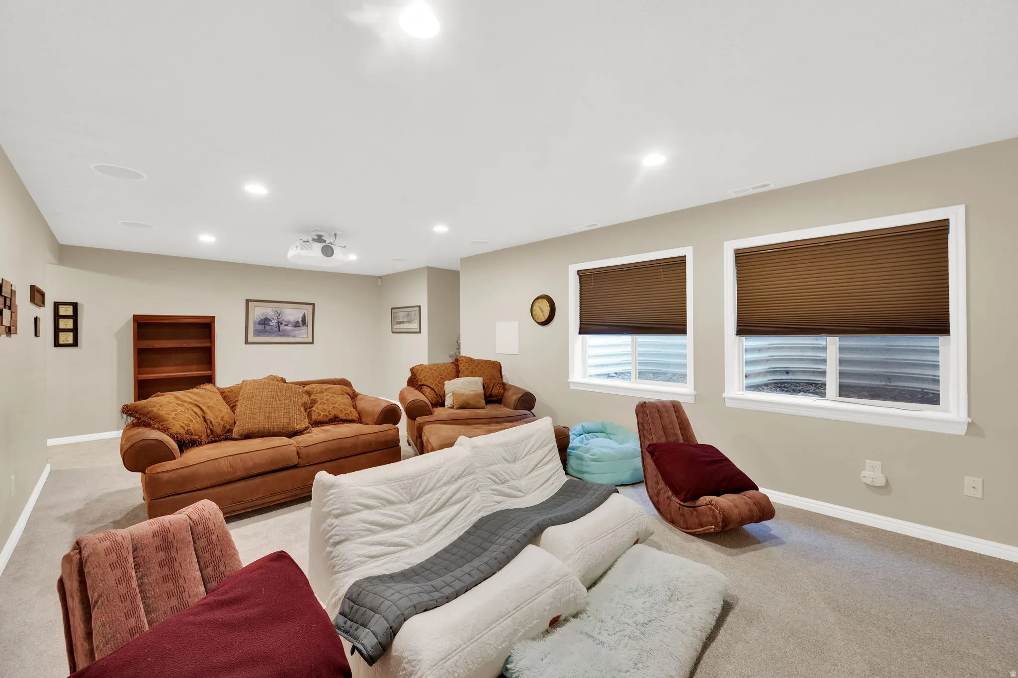 Spacious recreation area featuring recessed lighting, light neutral wall tones, two window wells with pleated shades, and wall-mounted speakers