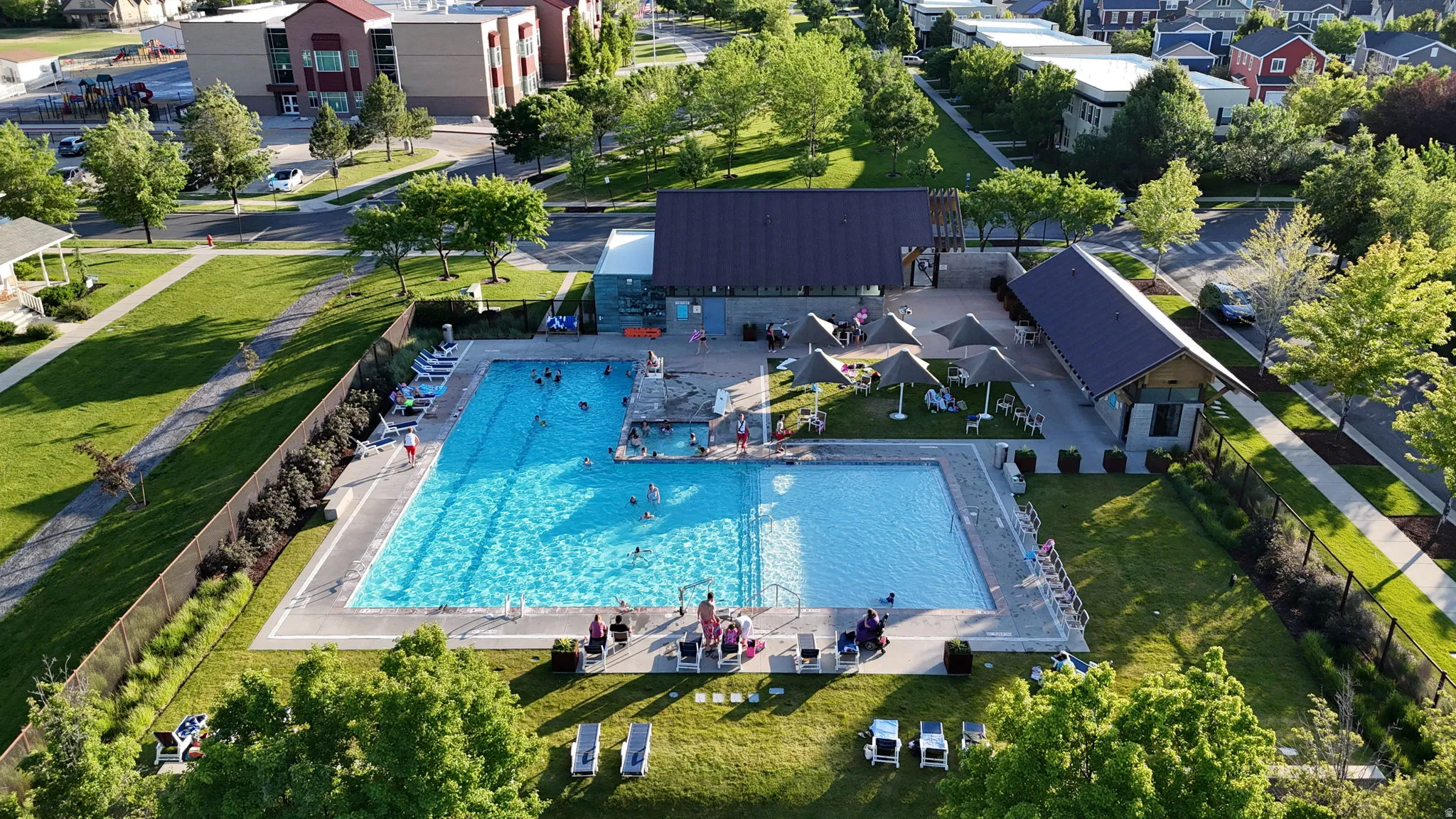 Community pool area featuring a large rectangular swimming pool, an adjacent smaller pool, paved pool deck, and shaded lounge areas