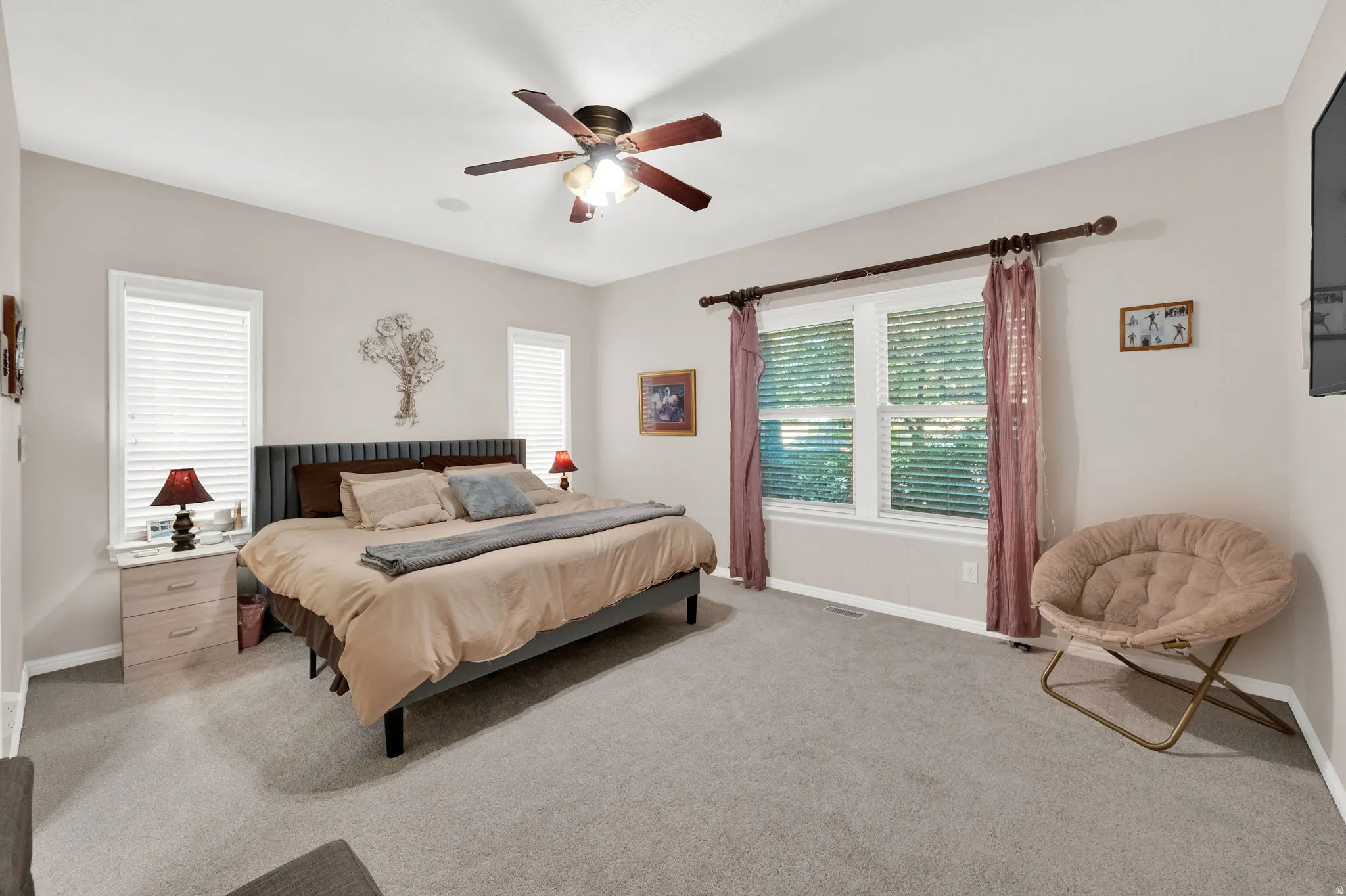 Spacious bedroom featuring plush carpet flooring, multiple windows with blinds, a ceiling fan with integrated lighting, and a wall-mounted television
