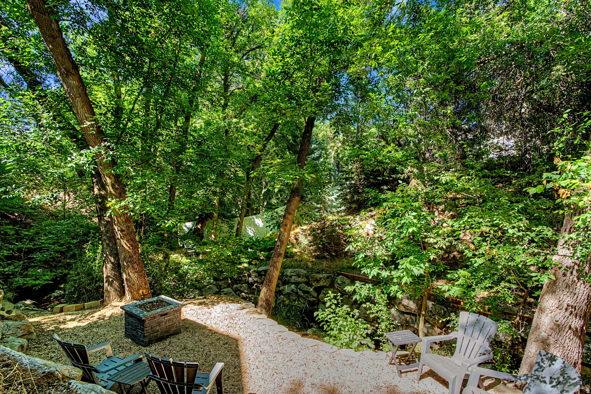 Secluded outdoor area featuring a gravel patio, mature trees, a built-in stone fire pit, and terraced landscaping