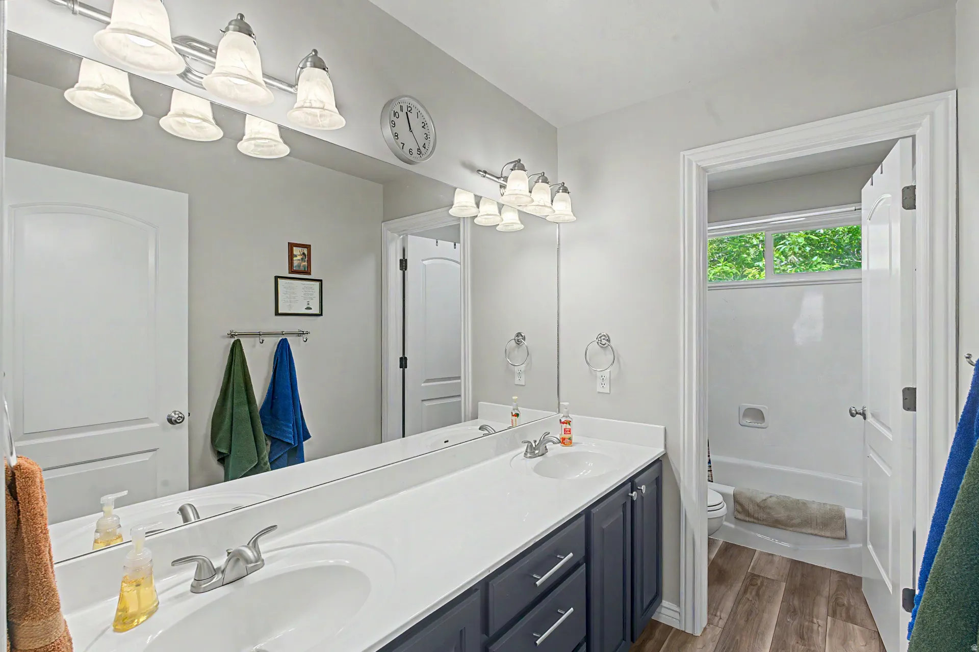 Bathroom featuring a dual vanity with a white countertop and dark cabinetry, wood-finish flooring, a full mirror, and a bathtub with an overhead window