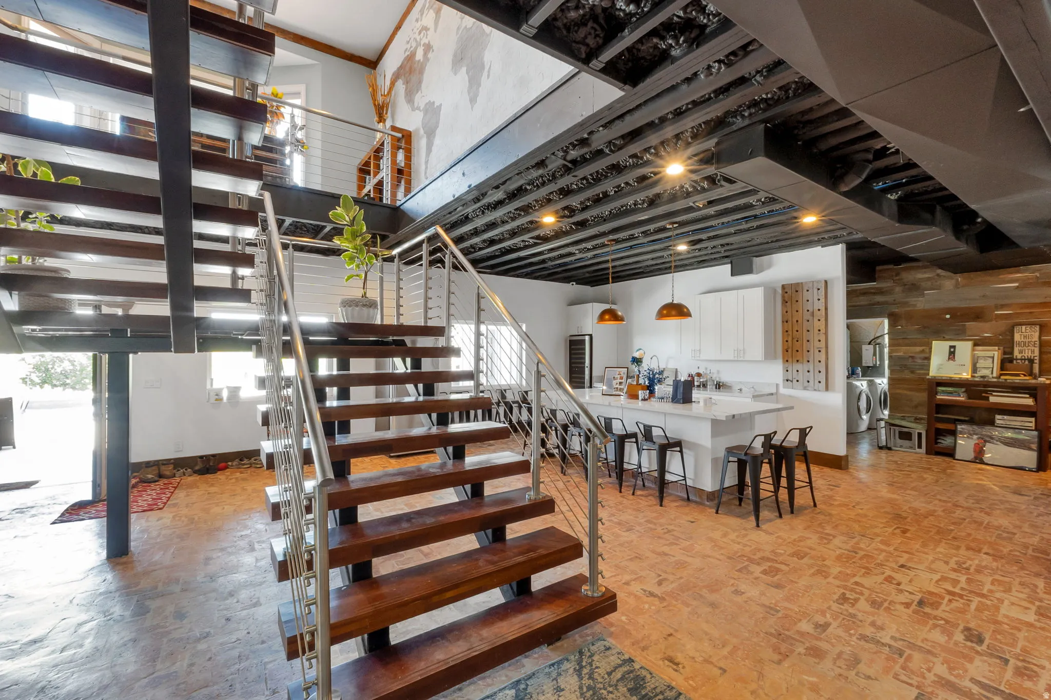 Open-concept living space featuring a floating staircase with wood treads and stainless steel railings, brick-finish flooring, a kitchen island with a white countertop and pendant lighting, white cabinetry, and a wood-paneled accent wall