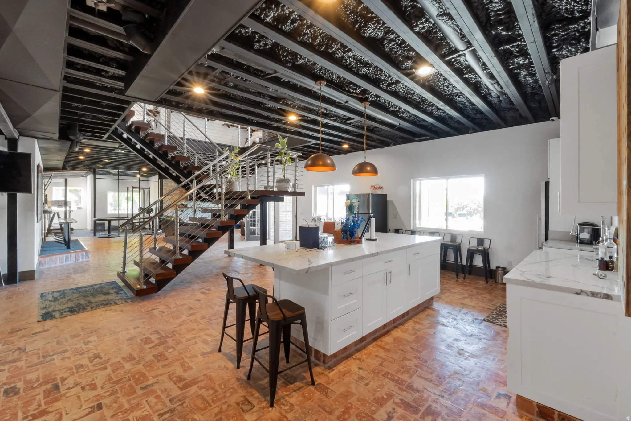 Open-concept living area featuring a central kitchen island with seating, a floating staircase with metal railings and wood treads, exposed ceiling beams with recessed lighting, and brick-finish flooring