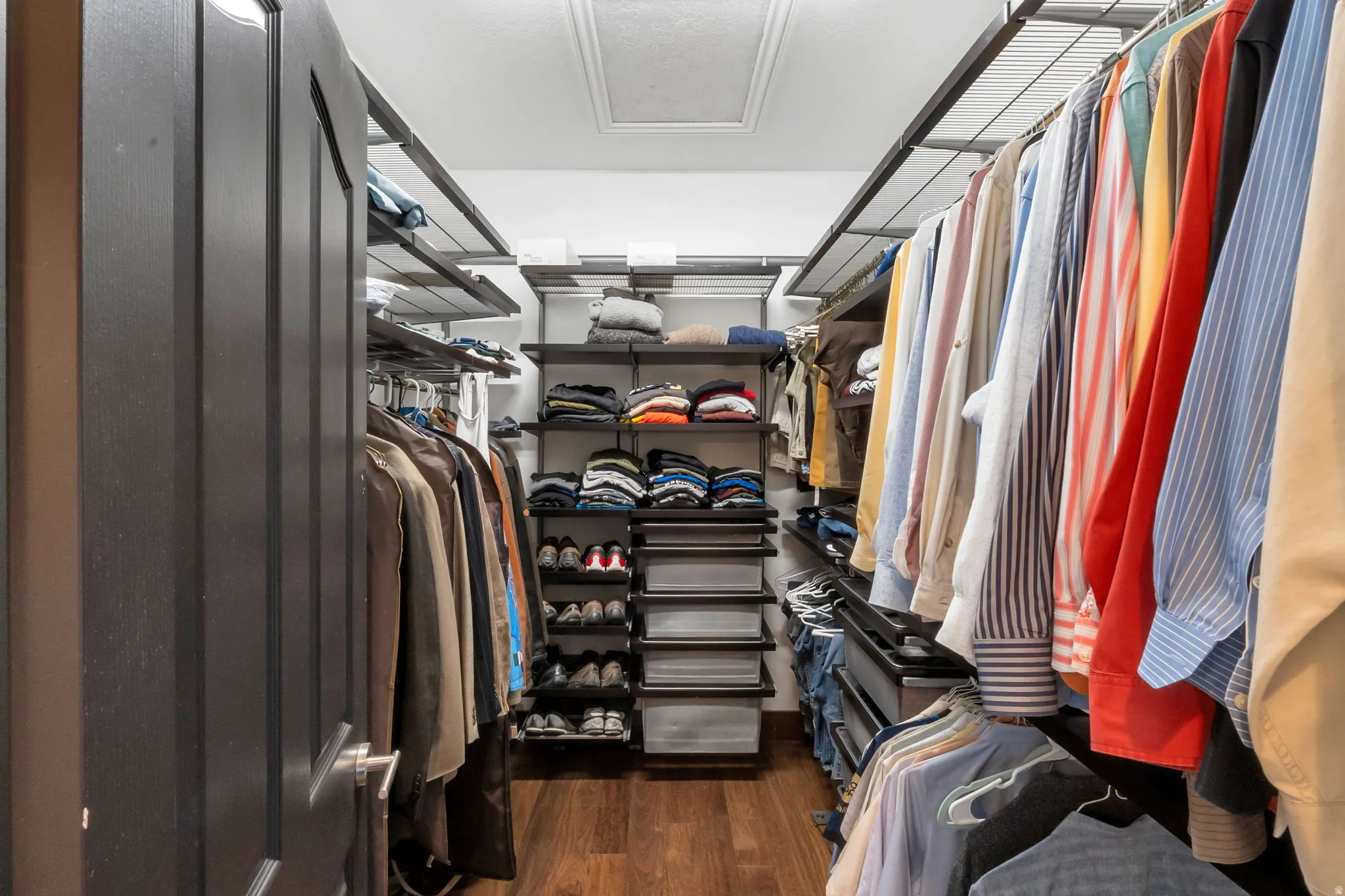 Walk-in closet featuring wood-finish flooring, dark panel door, multi-level shelving, integrated hanging rods, and an overhead attic access panel