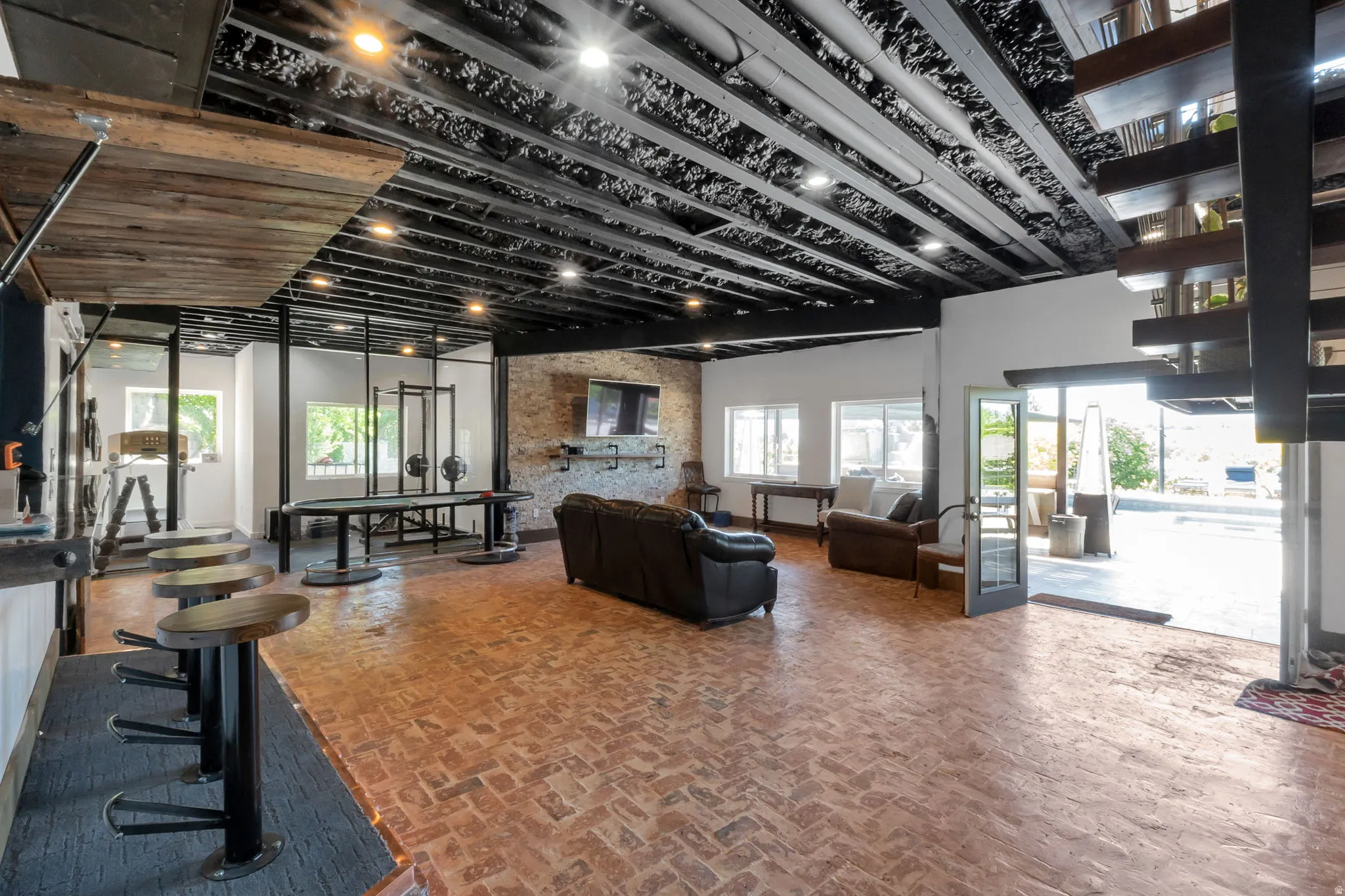 Spacious open-concept room featuring brick flooring, exposed ceiling beams, a built-in bar with seating, a prominent brick accent wall, and multiple windows