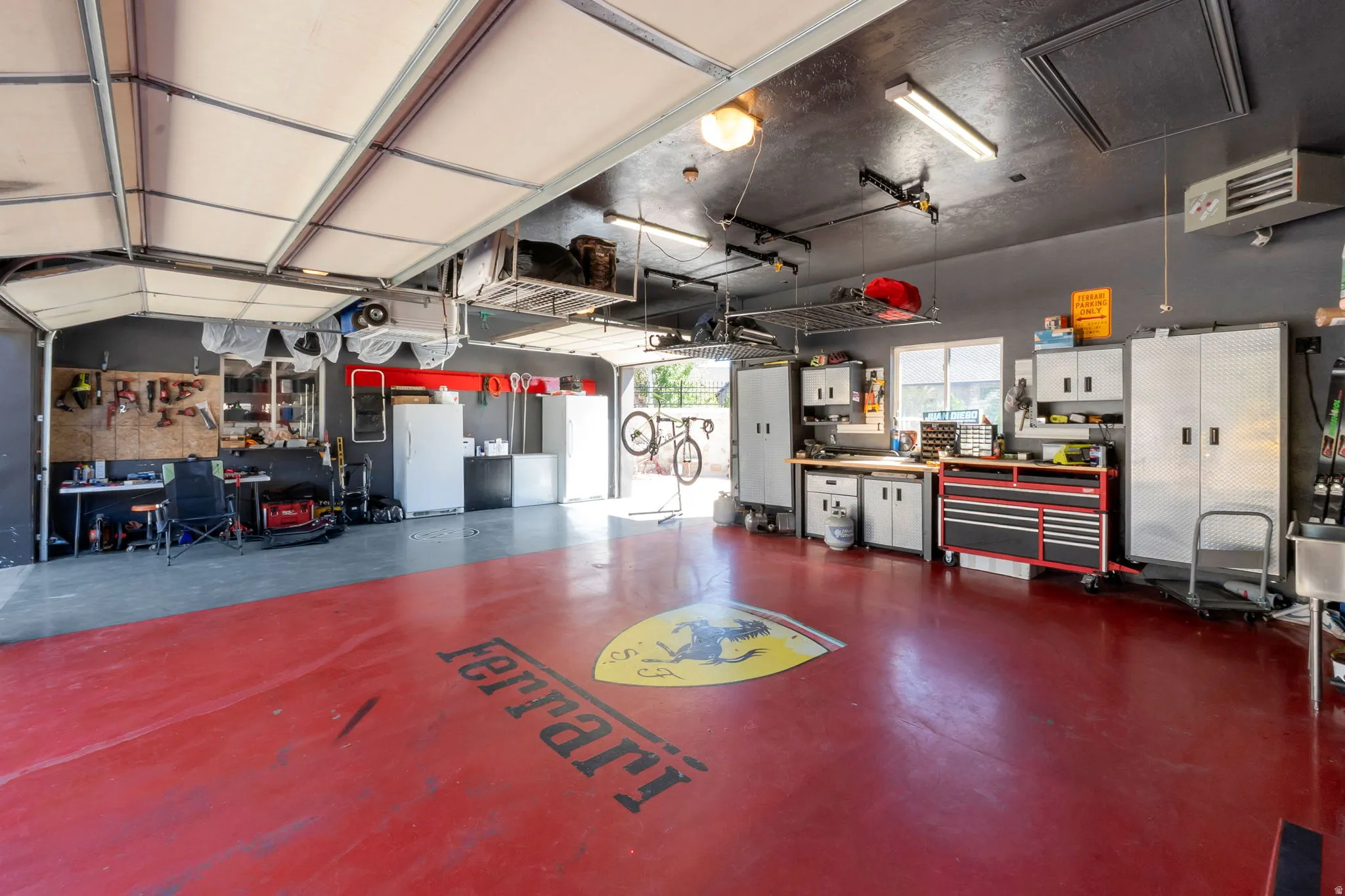 Spacious heated garage featuring a red concrete floor, overhead storage racks, fluorescent lighting, built-in cabinetry, and a utility sink