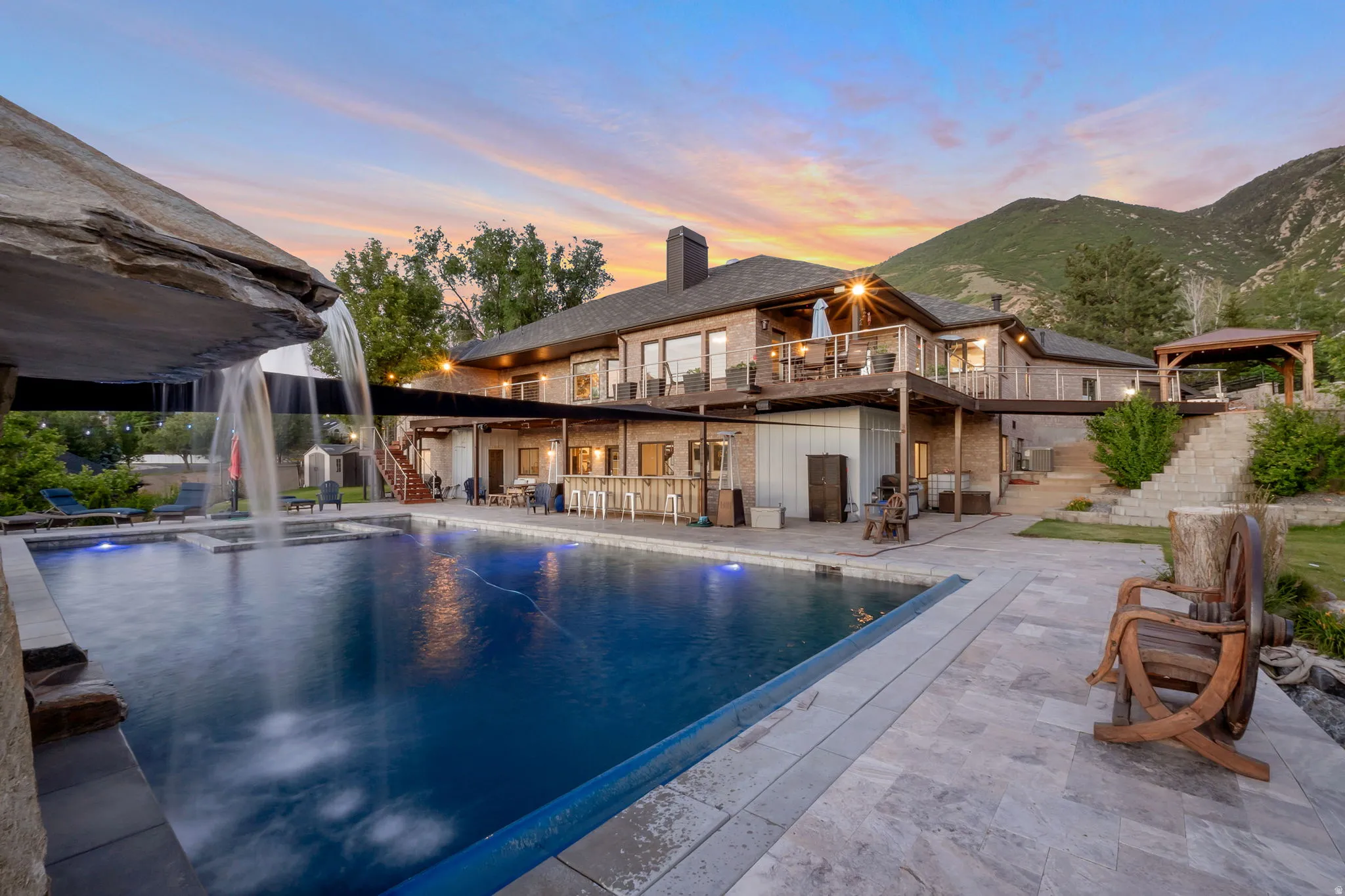 Resort-style outdoor area featuring a swimming pool with a rock waterfall, a spacious patio with light-toned pavers, a multi-level deck, an outdoor kitchen area, and a tiered staircase leading to an upper-level gazebo
