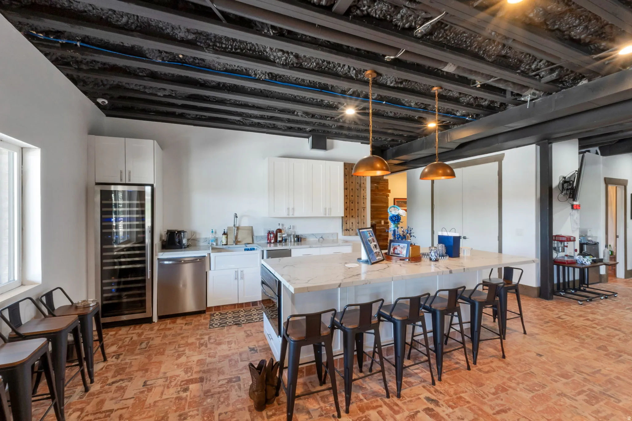 Kitchen space featuring an exposed ceiling with dark beams, brick-patterned flooring, white cabinetry, a large center island with a light-colored countertop, and a built-in wine refrigerator