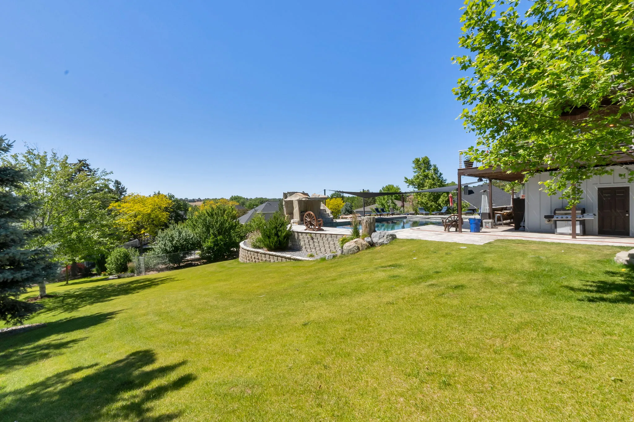 Expansive lawn area with a terraced retaining wall, surrounding mature trees, a swimming pool, and an outdoor patio featuring a pergola and a built-in outdoor kitchen with a grill station