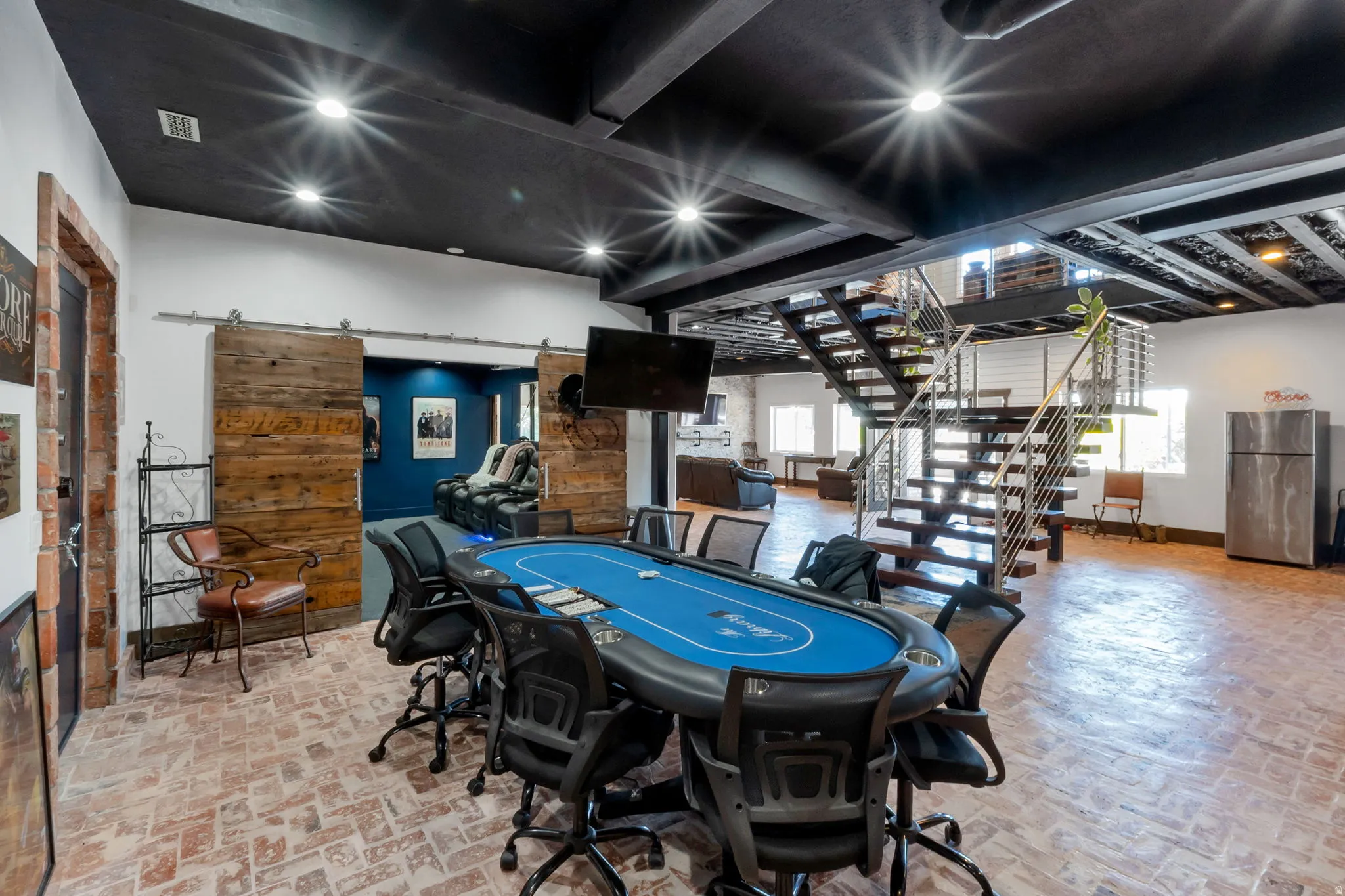 Open-concept recreation area featuring exposed brick flooring, a modern floating staircase with cable railing, and a black open-beam ceiling with recessed lighting