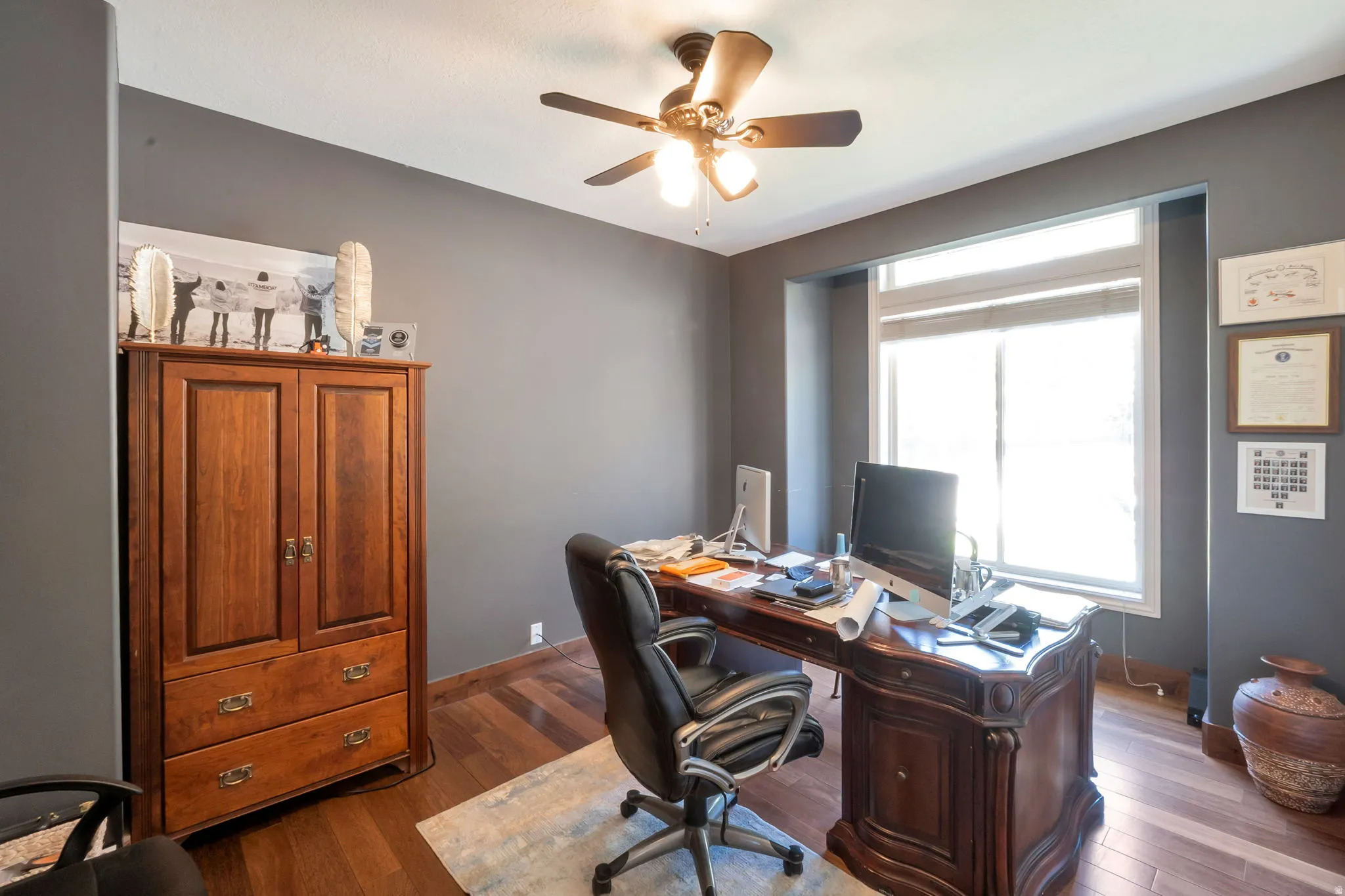 Room featuring dark gray walls, wood-finish flooring, a ceiling fan with light fixture, and a large window with white blinds