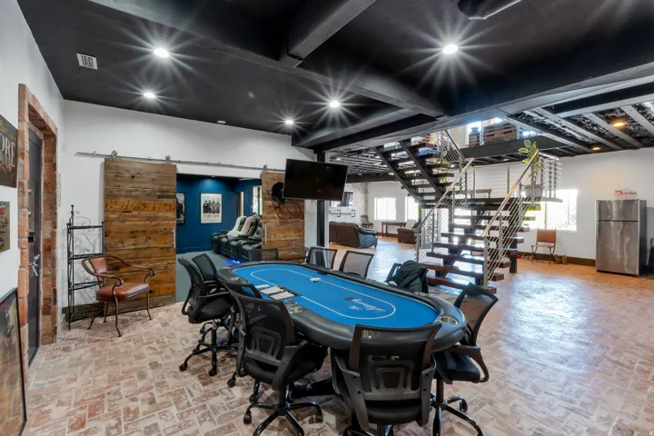Open-concept recreation area featuring exposed brick flooring, a modern floating staircase with cable railing, and a black open-beam ceiling with recessed lighting