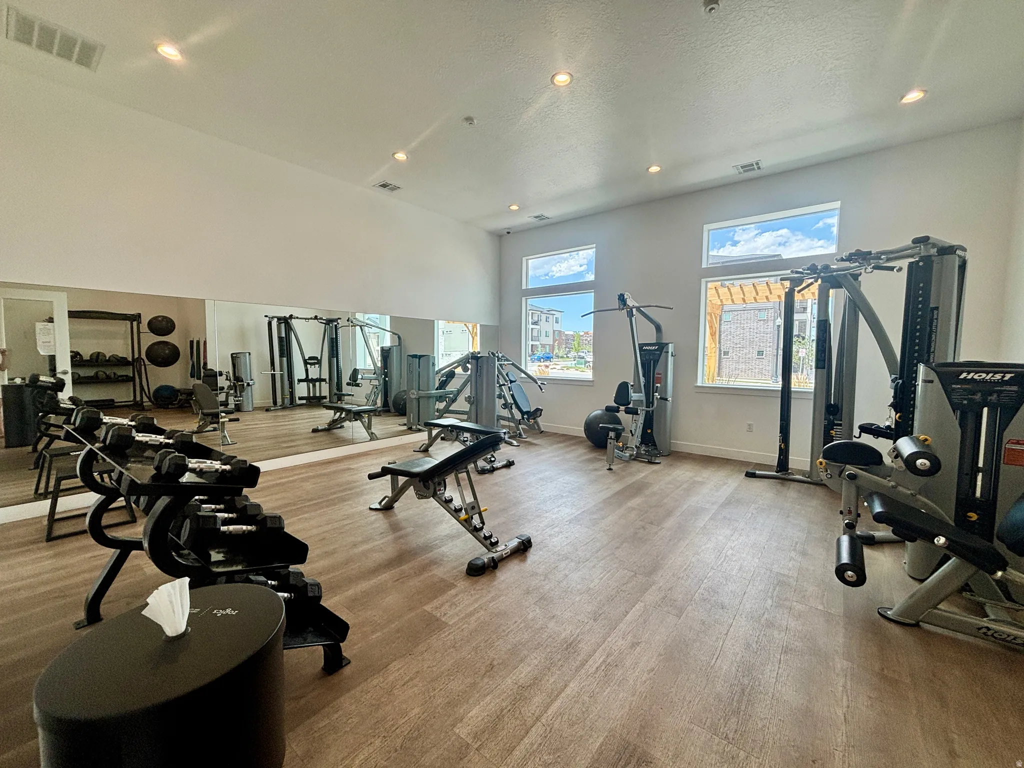 Spacious fitness center featuring wood-finish flooring, a full-length mirrored wall, multiple windows, recessed lighting, and various workout stations