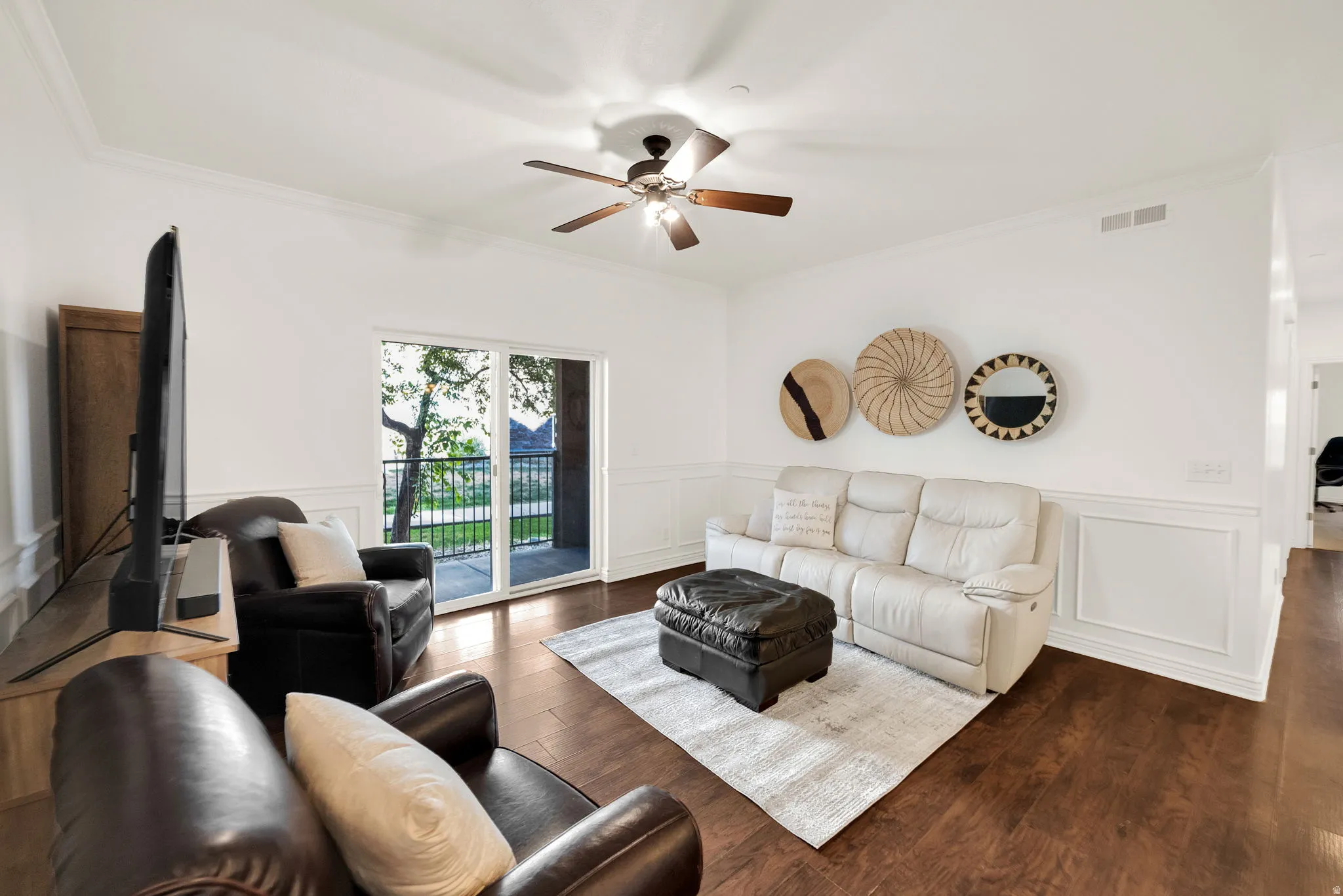 Living area featuring rich wood-finish flooring, white paneled wainscoting, crown molding, a ceiling fan with light fixture, and a sliding glass door opening to a private balcony