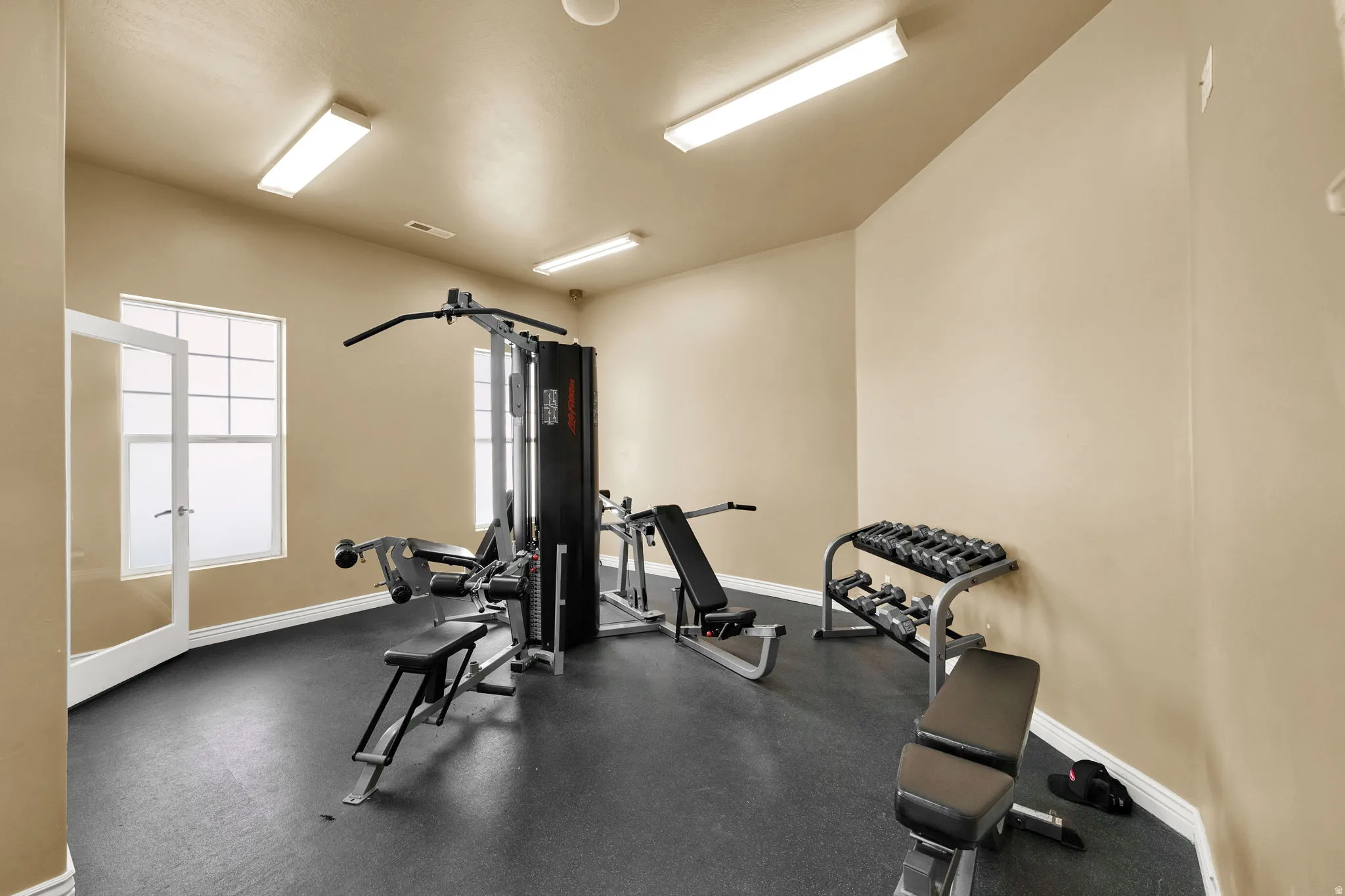 Dedicated exercise room featuring dark rubber flooring, two large windows with privacy coverings, and four overhead fluorescent light fixtures
