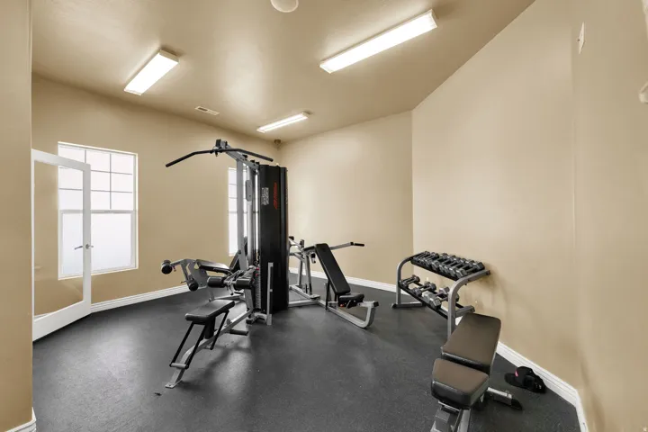 Dedicated exercise room featuring dark rubber flooring, two large windows with privacy coverings, and four overhead fluorescent light fixtures