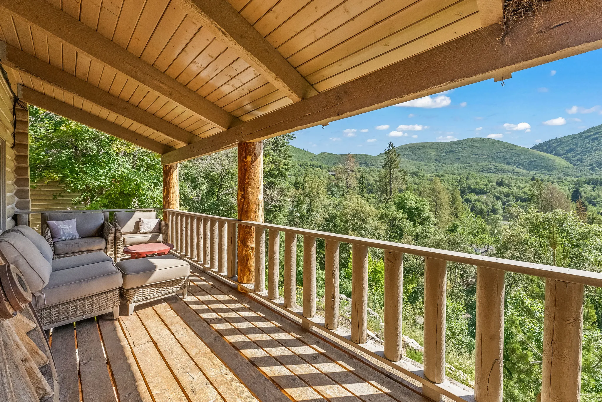 Covered outdoor deck featuring exposed wooden beams, natural wood railing, and plank flooring with extensive mountain and forest scenery