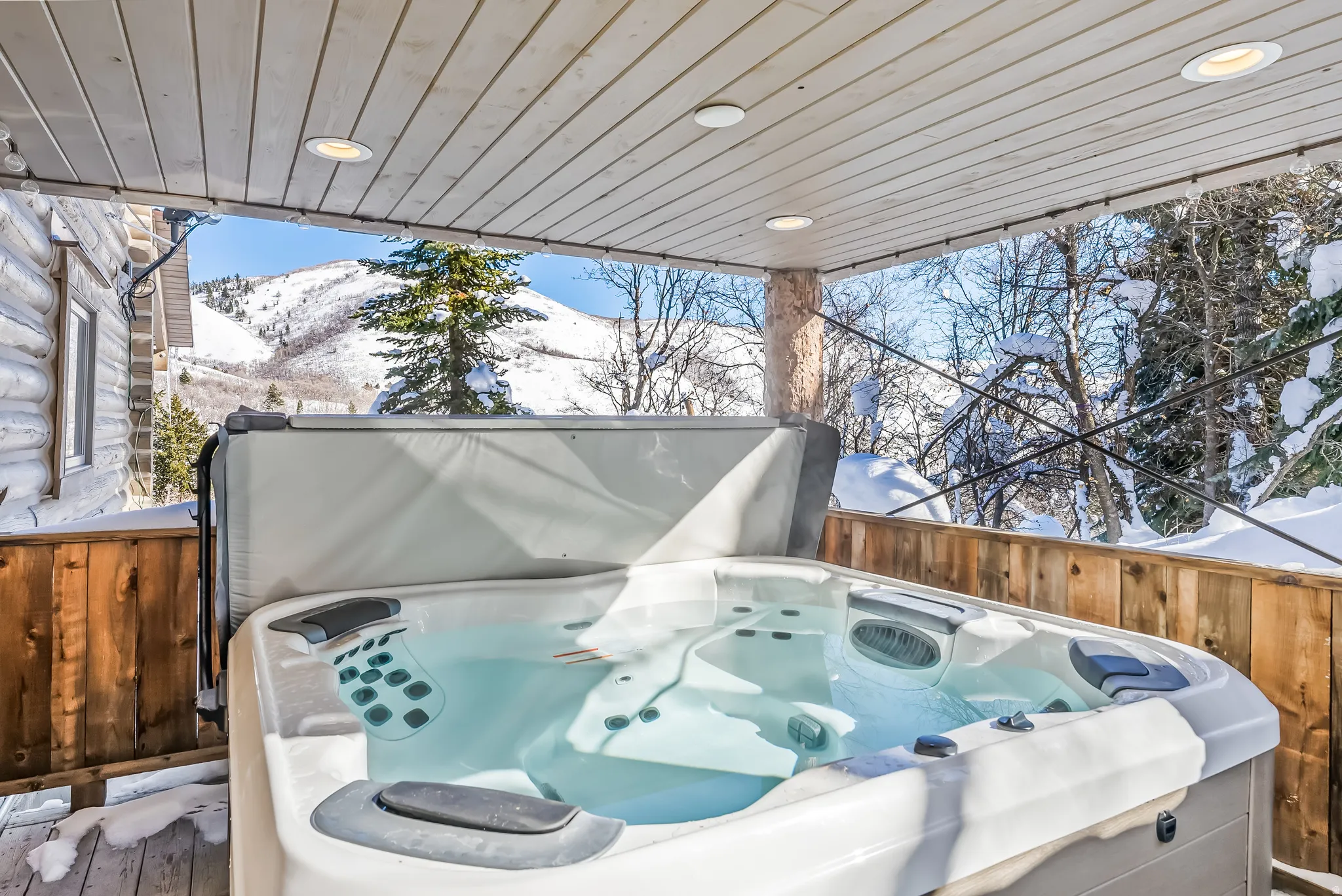 Covered outdoor hot tub with mountain scenery, log cabin exterior, wood-finish railing, and recessed ceiling lighting