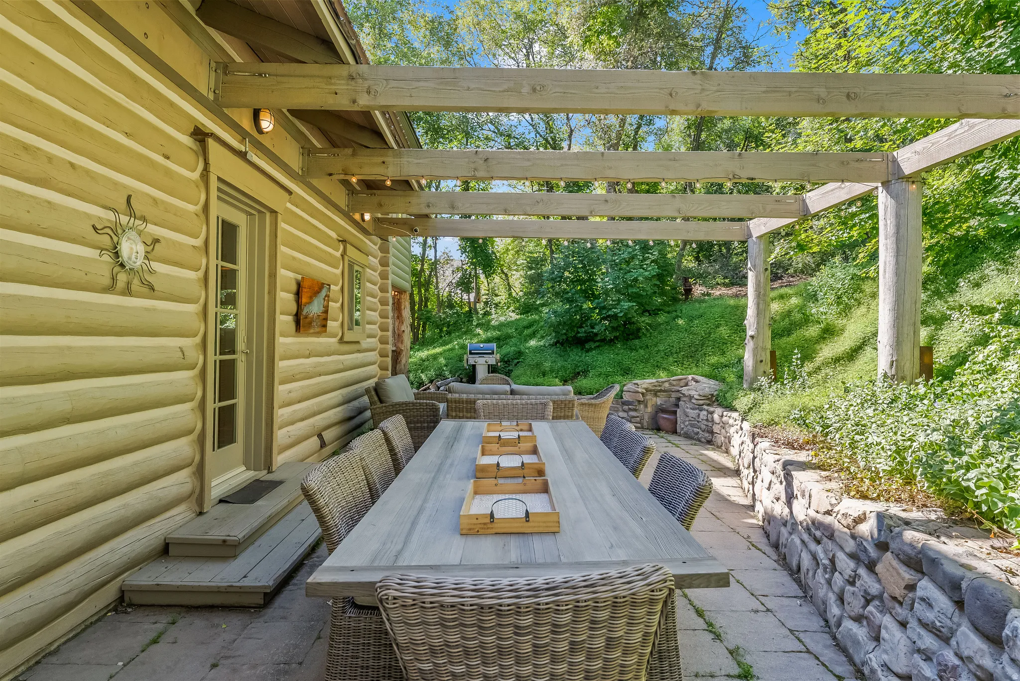 Expansive outdoor patio featuring a large dining area, wood pergola, stone retaining walls, and direct access to the home's log-style exterior