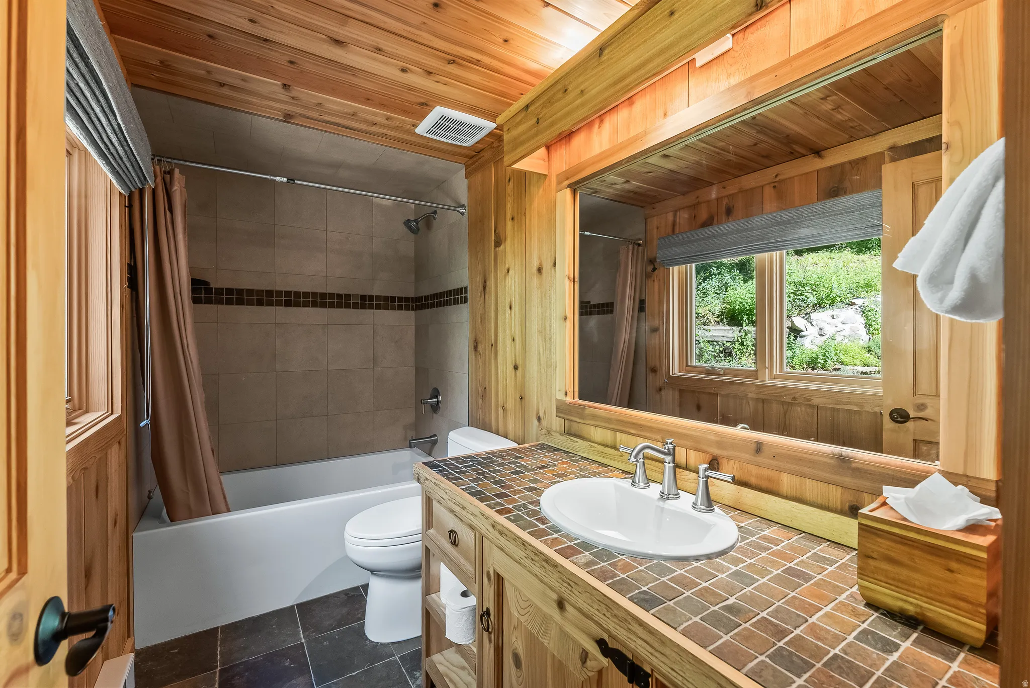 Bathroom featuring extensive wood paneling, a single vanity with a tile countertop, an oval undermount sink, and a framed mirror