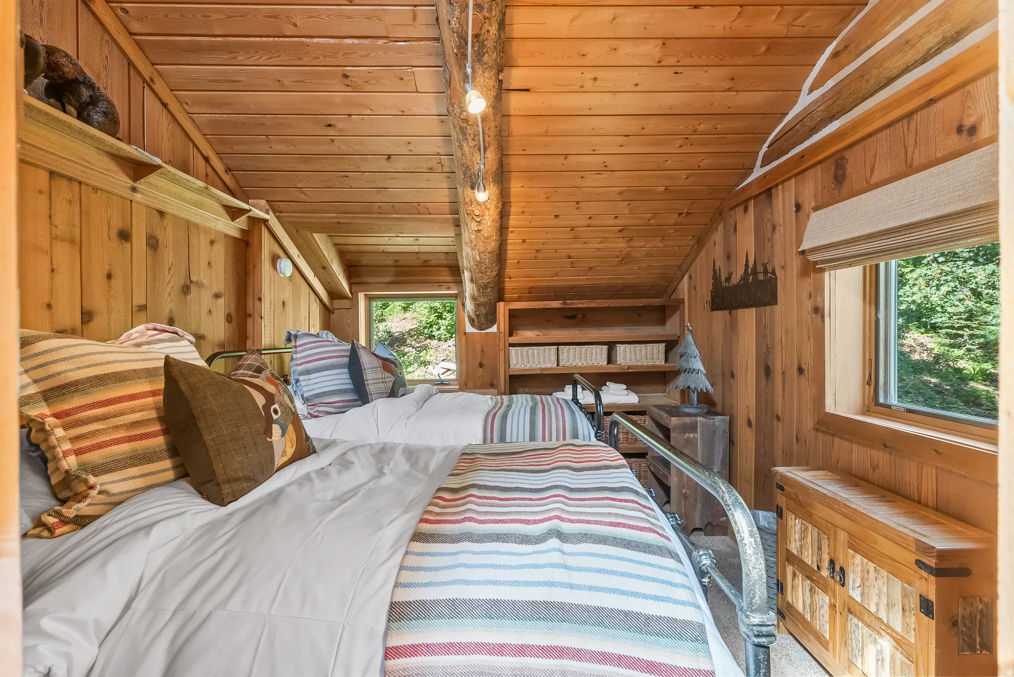 Wood paneling on walls and ceiling, exposed wooden beam, built-in shelving, two single beds with metal frames, and a window with a roller shade