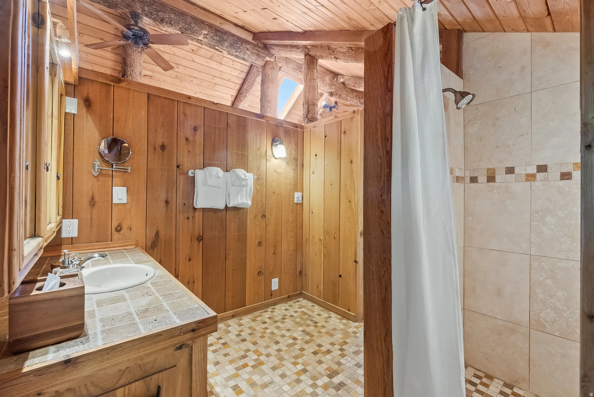 Rustic bathroom featuring wood-paneled walls and ceiling, a wood vanity with a tile countertop and integrated sink, and a tiled shower with an accent band