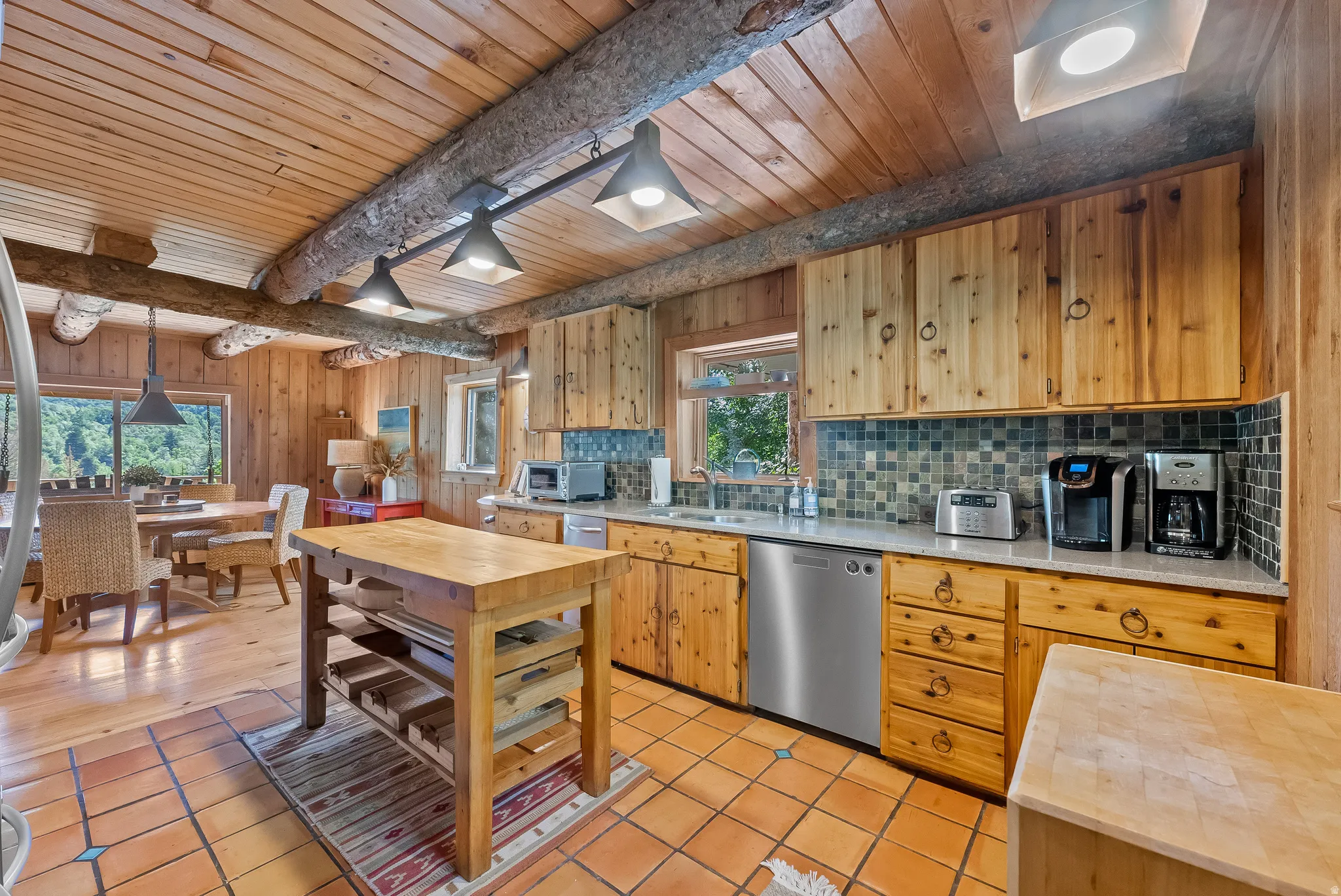 Wood-paneled kitchen featuring exposed log beams, custom wood cabinetry, stainless steel dishwasher, tile backsplash, and light-toned countertops