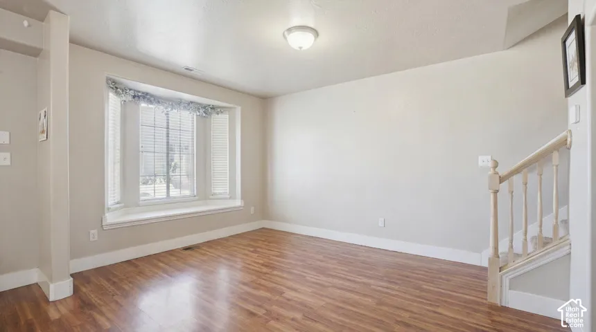 Spacious interior with wood-finish flooring, a prominent bay window with white blinds and trim, and a staircase featuring a wooden handrail and balusters