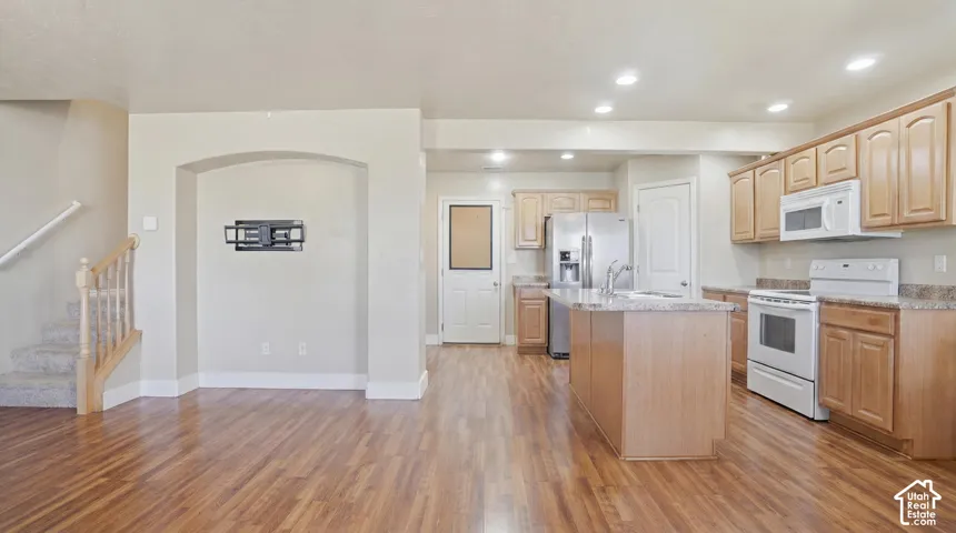 Open-concept kitchen with wood-finish flooring, light wood cabinetry, white appliances, and a central island featuring a sink and bar seating