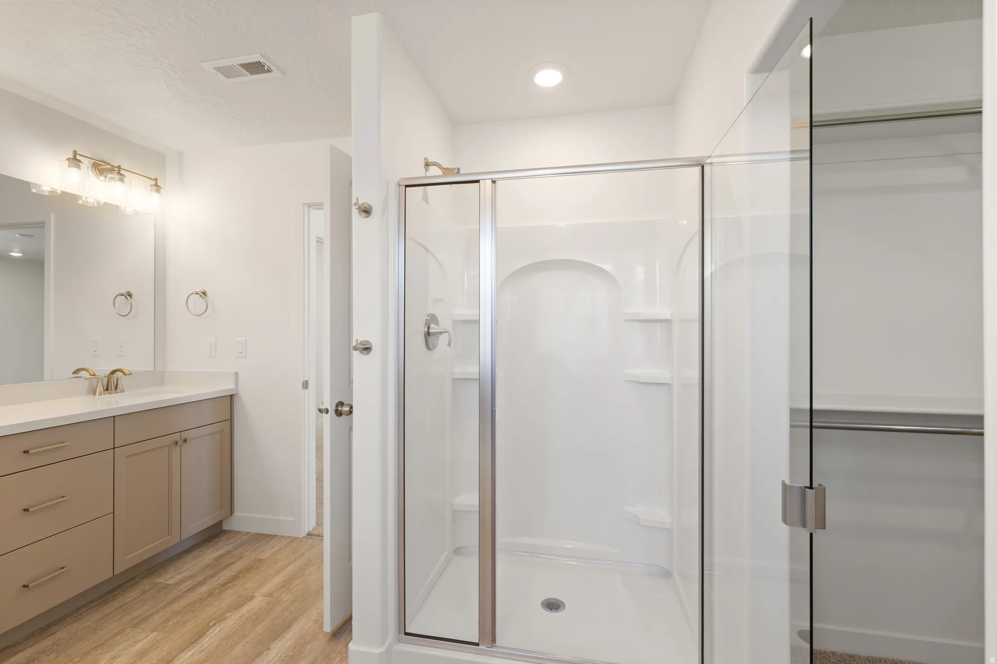 Contemporary bathroom featuring a walk-in shower with a glass enclosure, a vanity with light-toned cabinetry, light wood-finish flooring, a rectangular mirror, and an overhead vanity light fixture