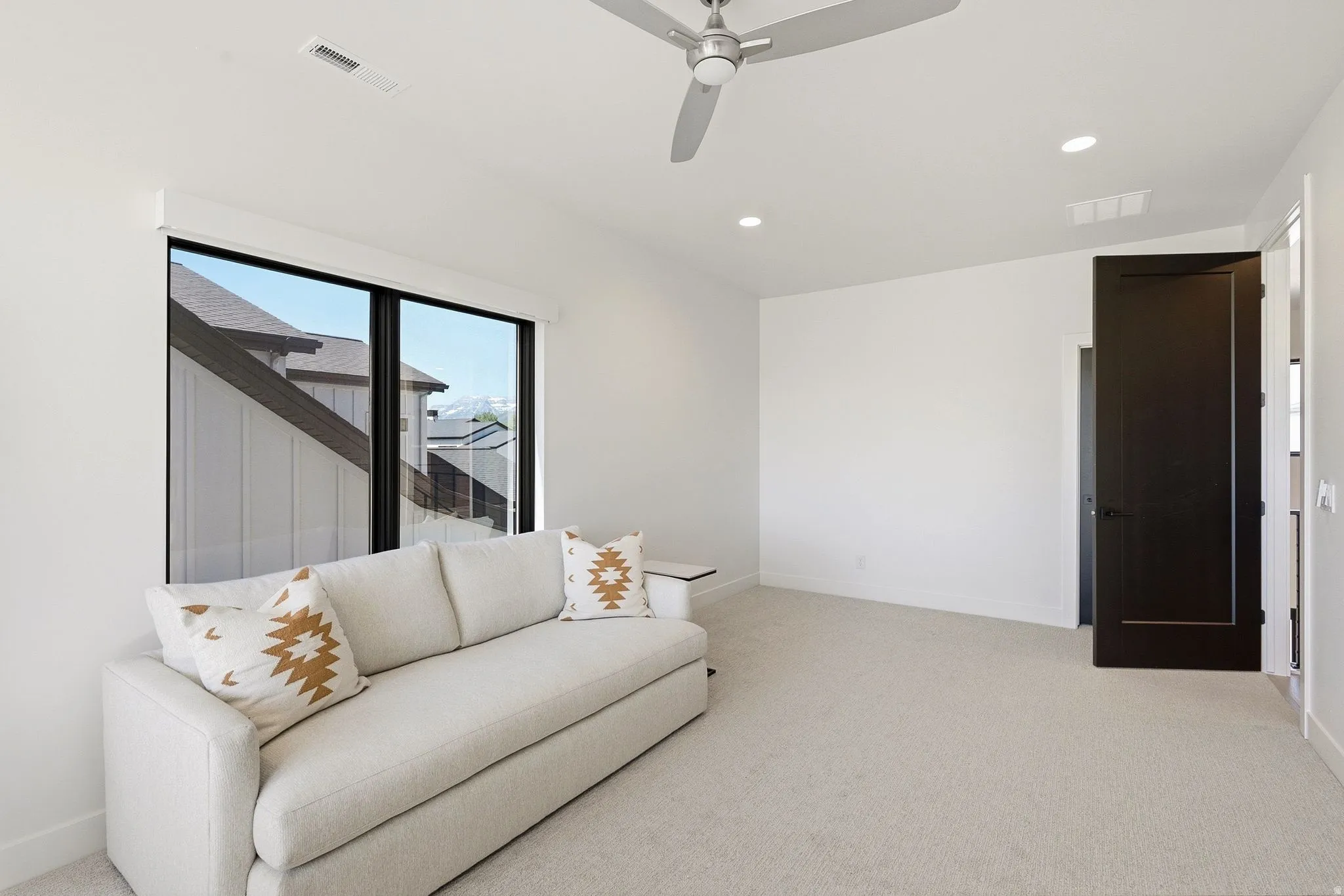 Bright room featuring a large window, light-toned carpet, recessed lighting, a ceiling fan, and a dark-paneled door
