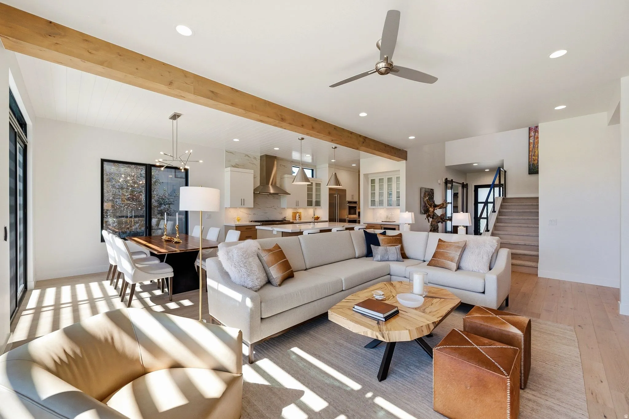 Spacious open-concept living area featuring exposed wood beams, light wood-finish flooring, a modern ceiling fan, recessed lighting, and a kitchen visible in the background