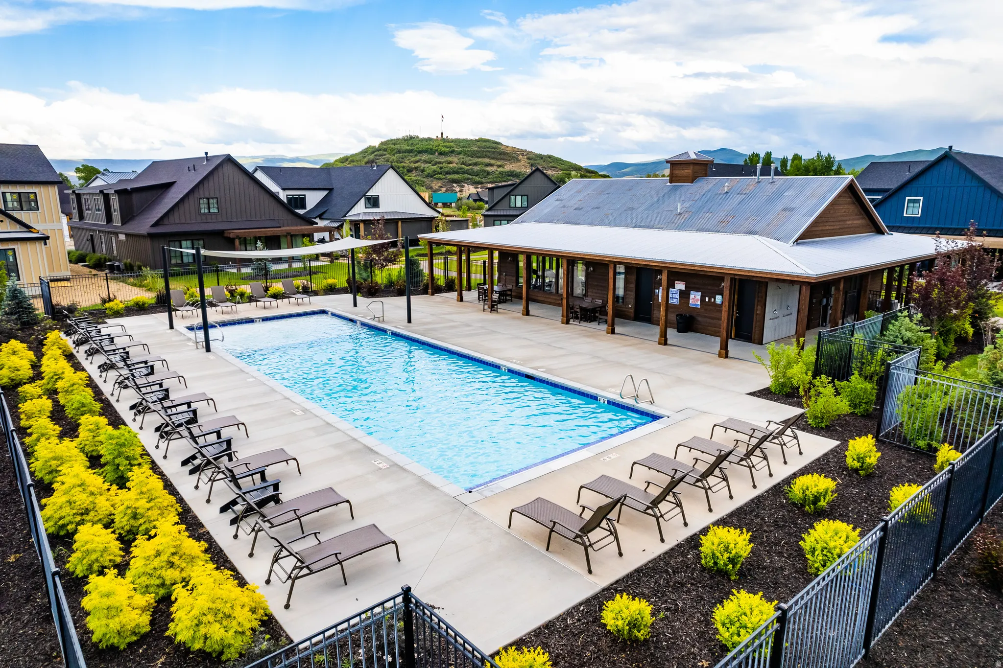 Community swimming pool with an expansive concrete deck, surrounded by black metal fencing and landscaped beds featuring vibrant green and yellow shrubbery