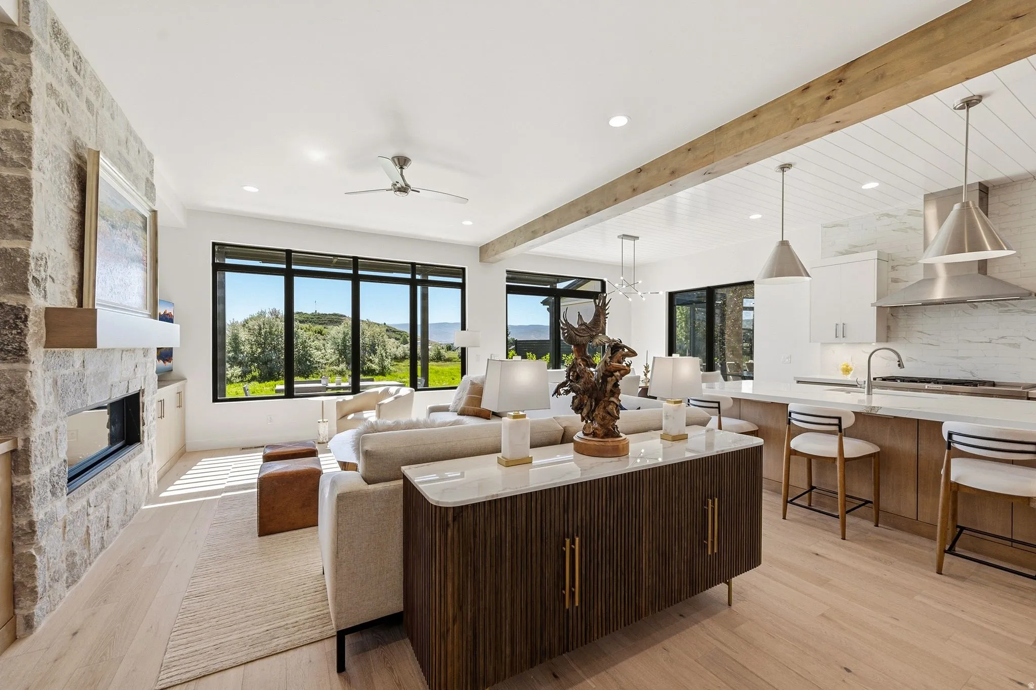 Open-concept living space featuring expansive windows, light wood-finish flooring, a stone-clad fireplace with a built-in hearth, a kitchen island with seating, and a decorative wood beam