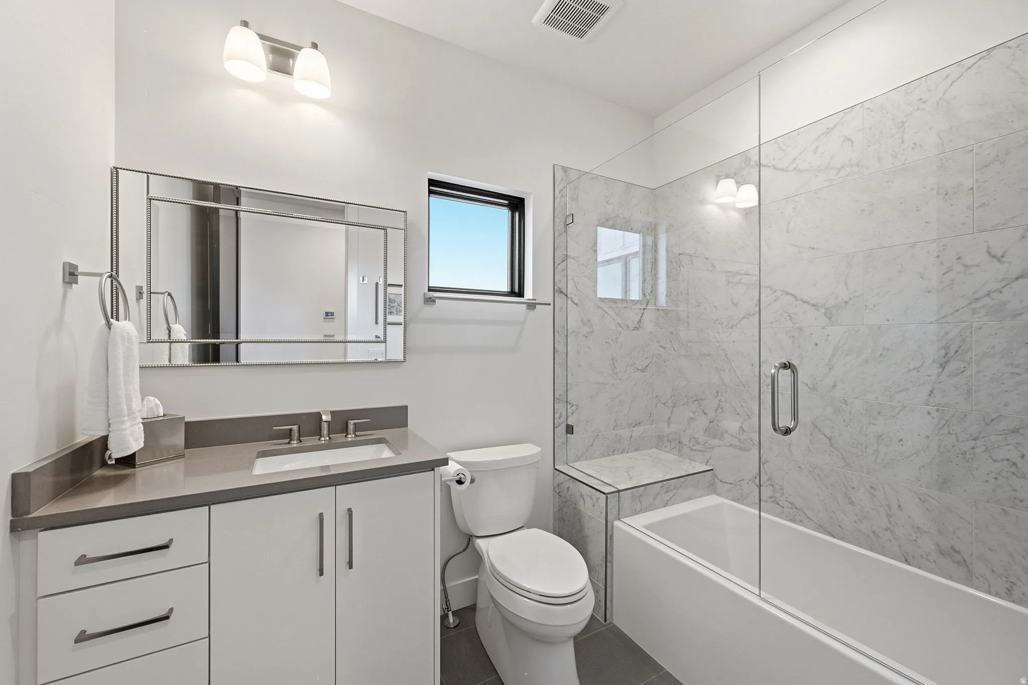 Full bathroom featuring a rectangular vanity mirror, a single vanity with a gray countertop and undermount sink, and a combination tub/shower with a glass enclosure and marble-look tile surround