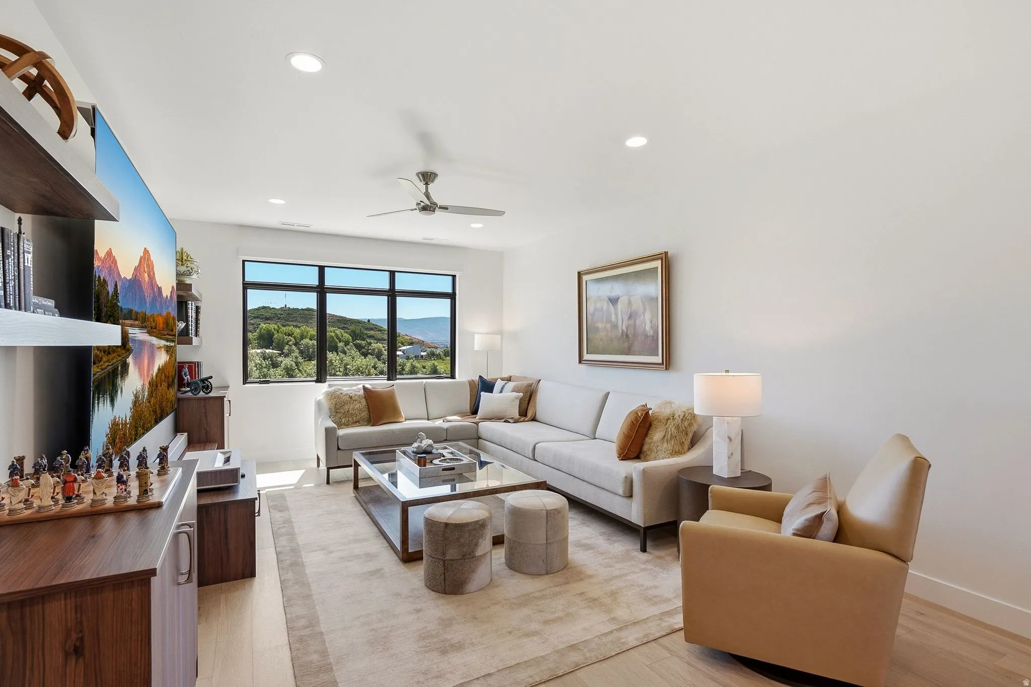Spacious living area featuring large windows showcasing surrounding scenery, light wood-finish flooring, recessed lighting, and a ceiling fan